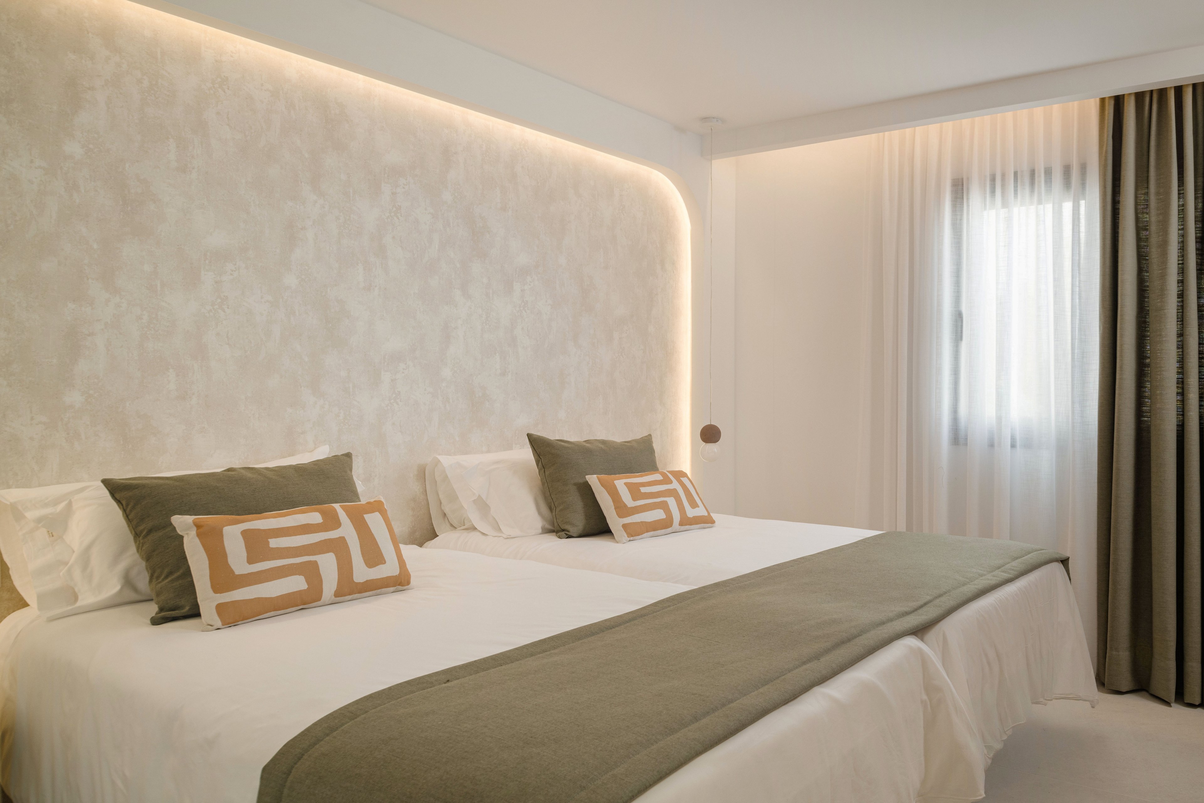 Contemporary hotel room featuring two neatly made twin beds with white linens and green throws. The headboard wall has soft textured finish and integrated warm LED strip lighting that creates a cozy ambiance. Decorative pillows with geometric patterns add visual interest, while sheer curtains and a side drape frame the window. The overall design is minimal, bright, and inviting, emphasizing comfort and relaxation.