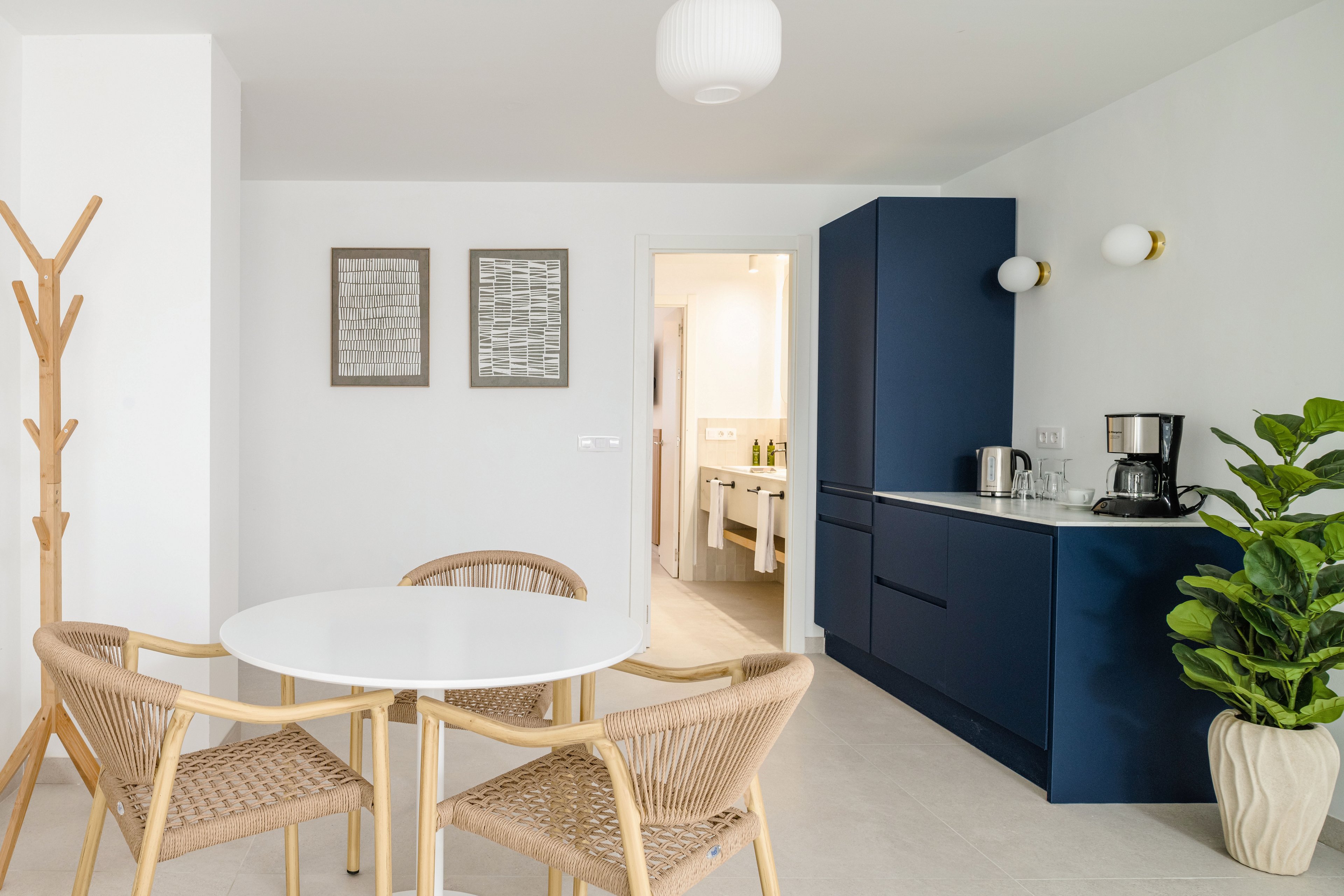 Bright contemporary kitchen space features a round white dining table with woven chairs and a sleek blue cabinet unit. Small appliances, glassware, and a potted plant add functional and decorative elements to the room. A wooden coat rack and framed wall art enhance the minimalist aesthetic. The open doorway reveals an adjacent room with additional storage and bottles, extending the clean, modern design.