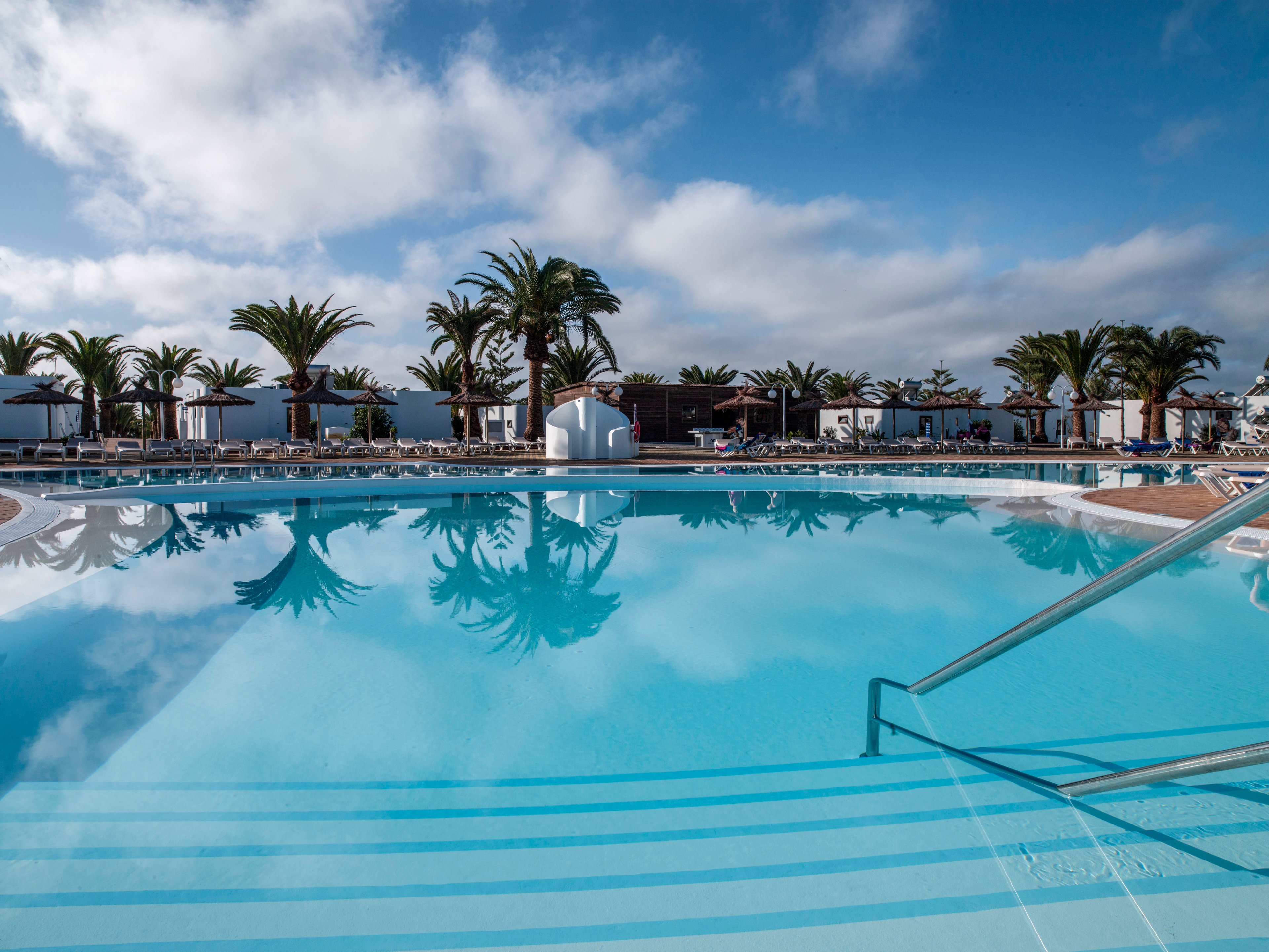 A large, clear blue swimming pool is the central focus of this resort scene. Palm trees and white low-rise buildings line the background, reflected in the calm water. Sun loungers and umbrellas are arranged around the pool, suggesting a relaxing holiday atmosphere. The sky is partly cloudy with bright sunlight, enhancing the tranquil, inviting mood.