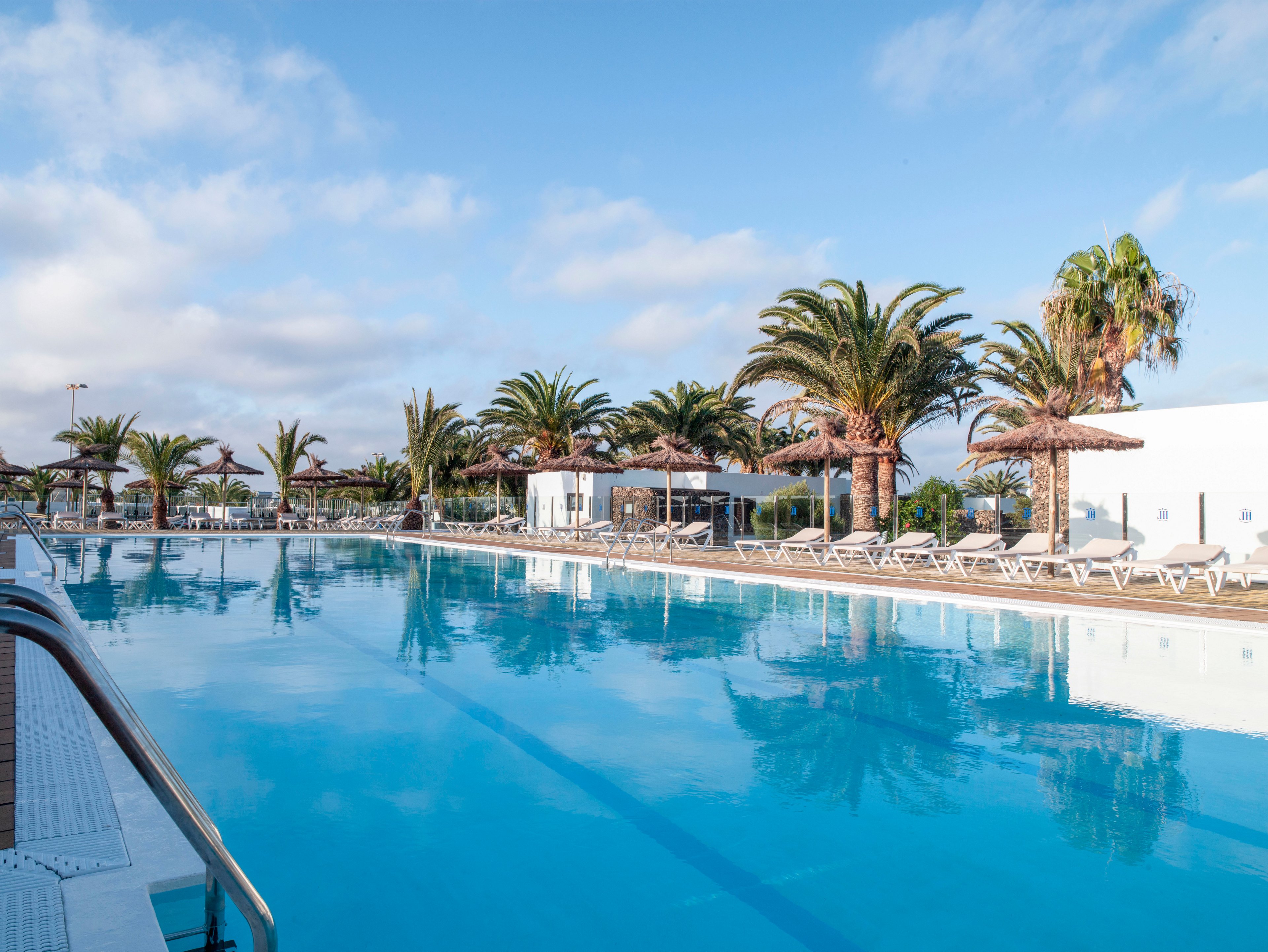 A large outdoor swimming pool reflects the blue sky on a clear sunny day. Palm trees and straw parasols line the poolside, creating a relaxed vacation atmosphere. White lounge chairs are neatly arranged along the deck, suggesting a quiet resort setting. Bright natural light and soft clouds enhance the tranquil, inviting mood of the scene.