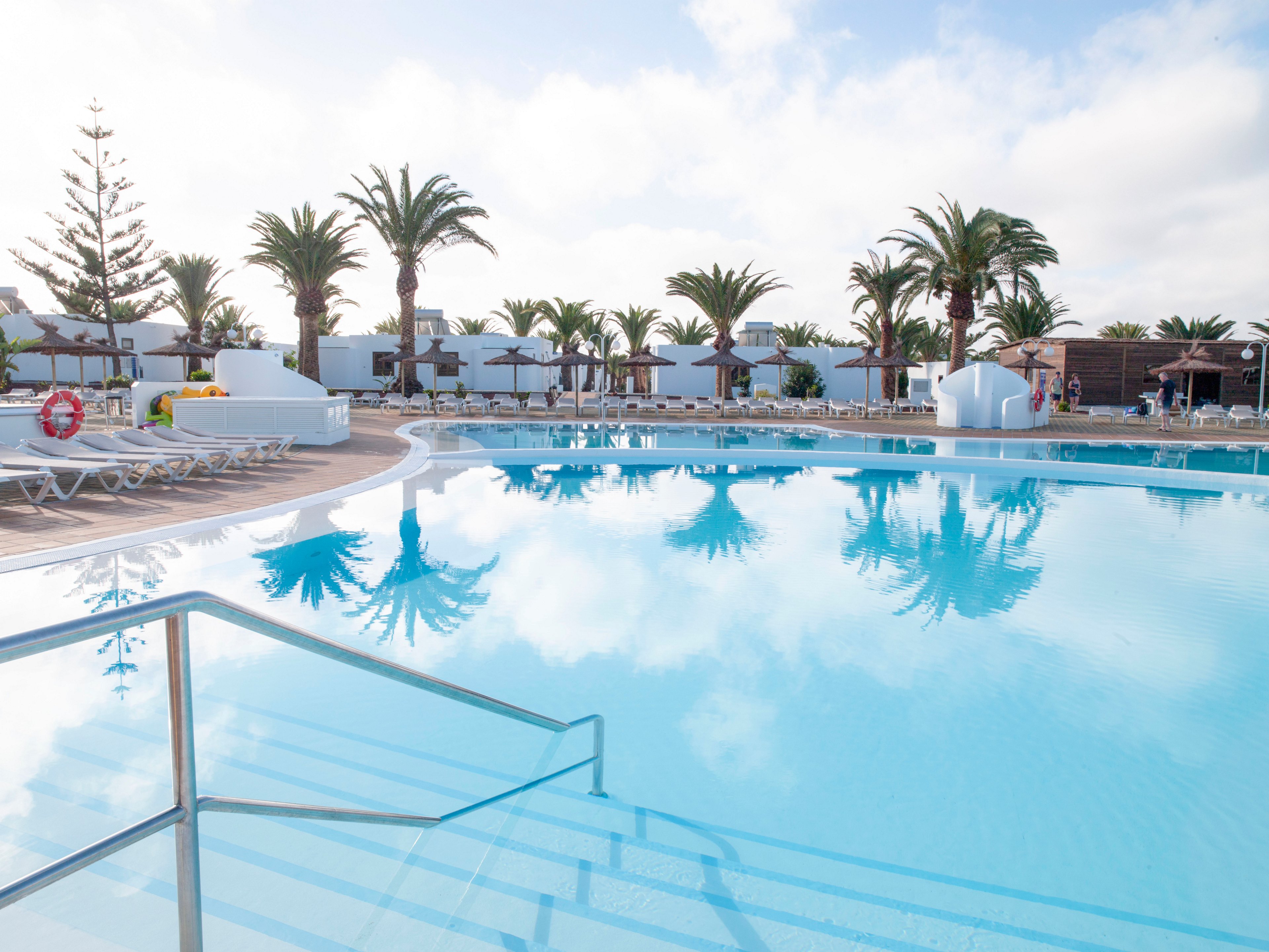 A large, clear swimming pool reflects palm trees and white resort buildings under a bright sky. Sun loungers line the poolside, suggesting a relaxing vacation setting. Stainless steel handrails lead into the shallow end of the pool, emphasizing easy access for swimmers. The overall scene feels calm, spacious, and inviting for holidaymakers.