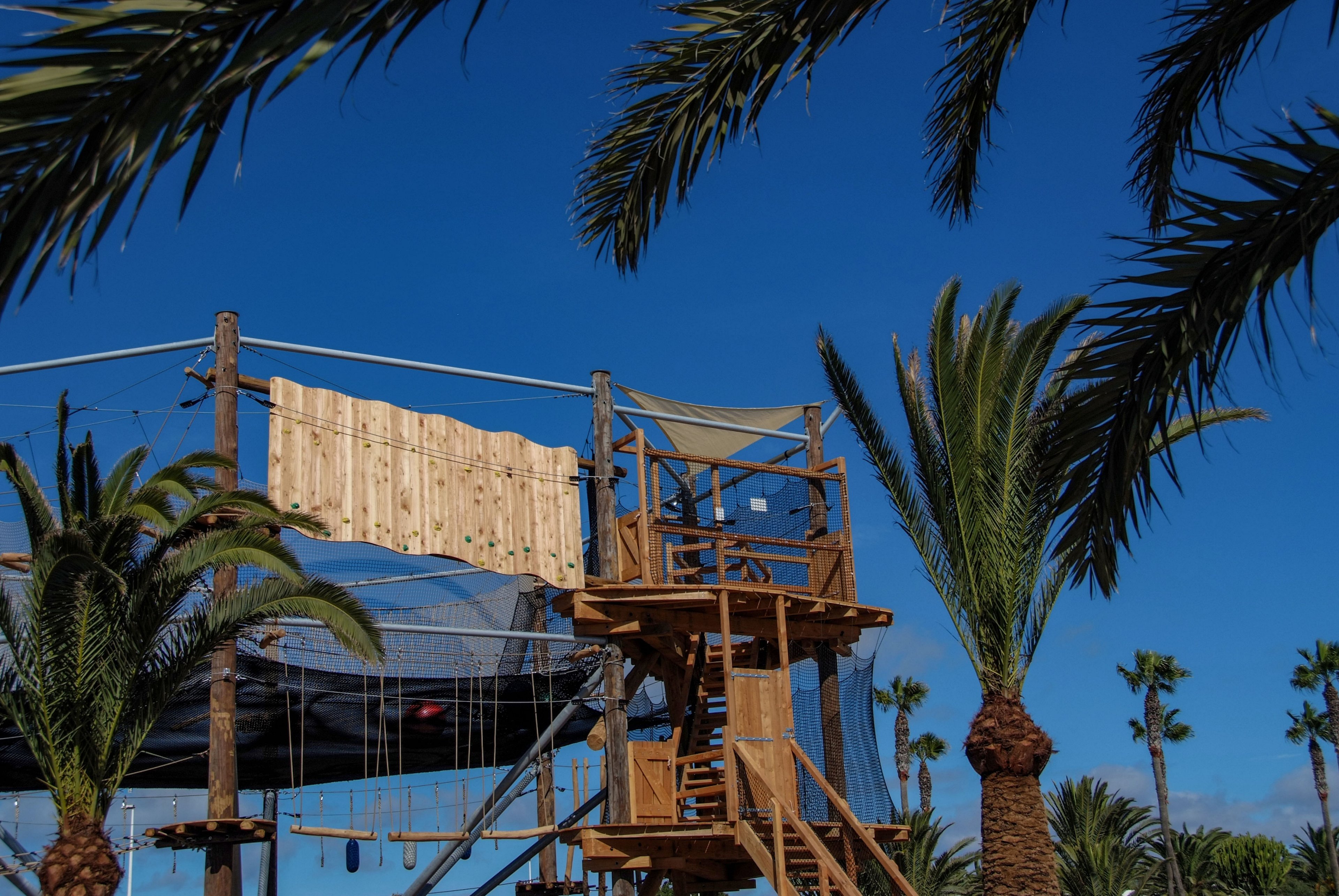 An elevated wooden ropes course structure stands among tall palm trees under a clear blue sky. The adventure park features platforms, climbing elements, and suspended ropes and nets. Sunlight highlights the warm tones of the wood against the vibrant greenery. The scene suggests a recreational outdoor activity area in a warm, coastal or tropical environment.