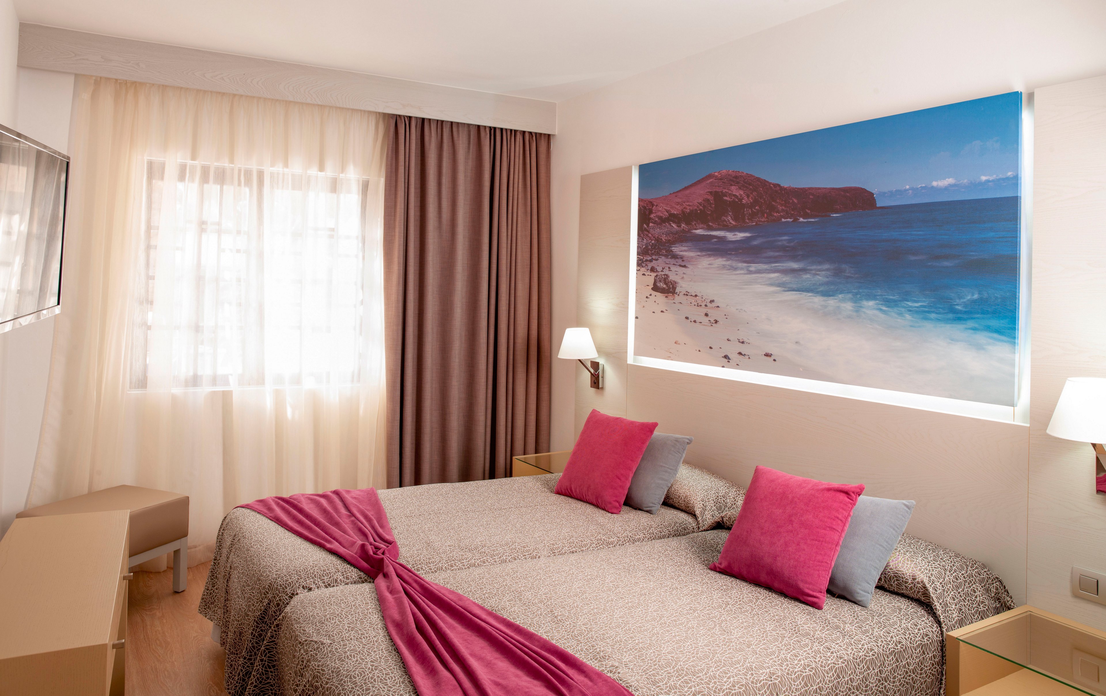 A cozy twin hotel room features two single beds with patterned bedspreads and bright pink accent pillows. A large illuminated wall print of a beach landscape adds a coastal theme to the space. Soft natural light enters through a window with sheer curtains, complemented by darker drapes. Neutral furniture and bedside lamps create a clean, modern and inviting atmosphere.