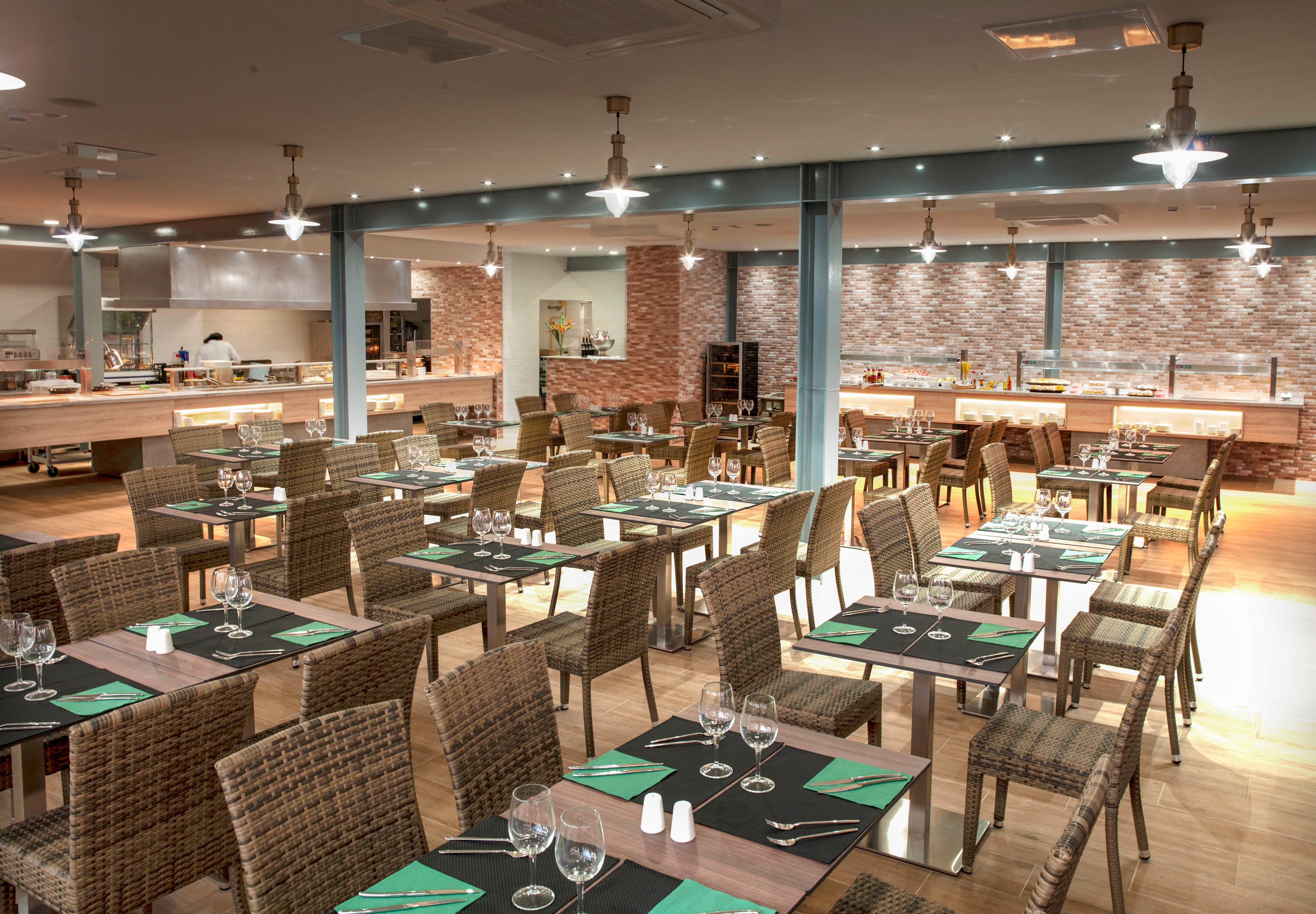 A contemporary restaurant dining area features rows of wicker chairs and wooden tables neatly set with glassware and cutlery. Green napkins and white salt and pepper shakers add accents to the neutral interior. In the background, a buffet counter and open kitchen are visible against a brick wall. Warm lighting from ceiling fixtures creates an inviting atmosphere in the spacious room.