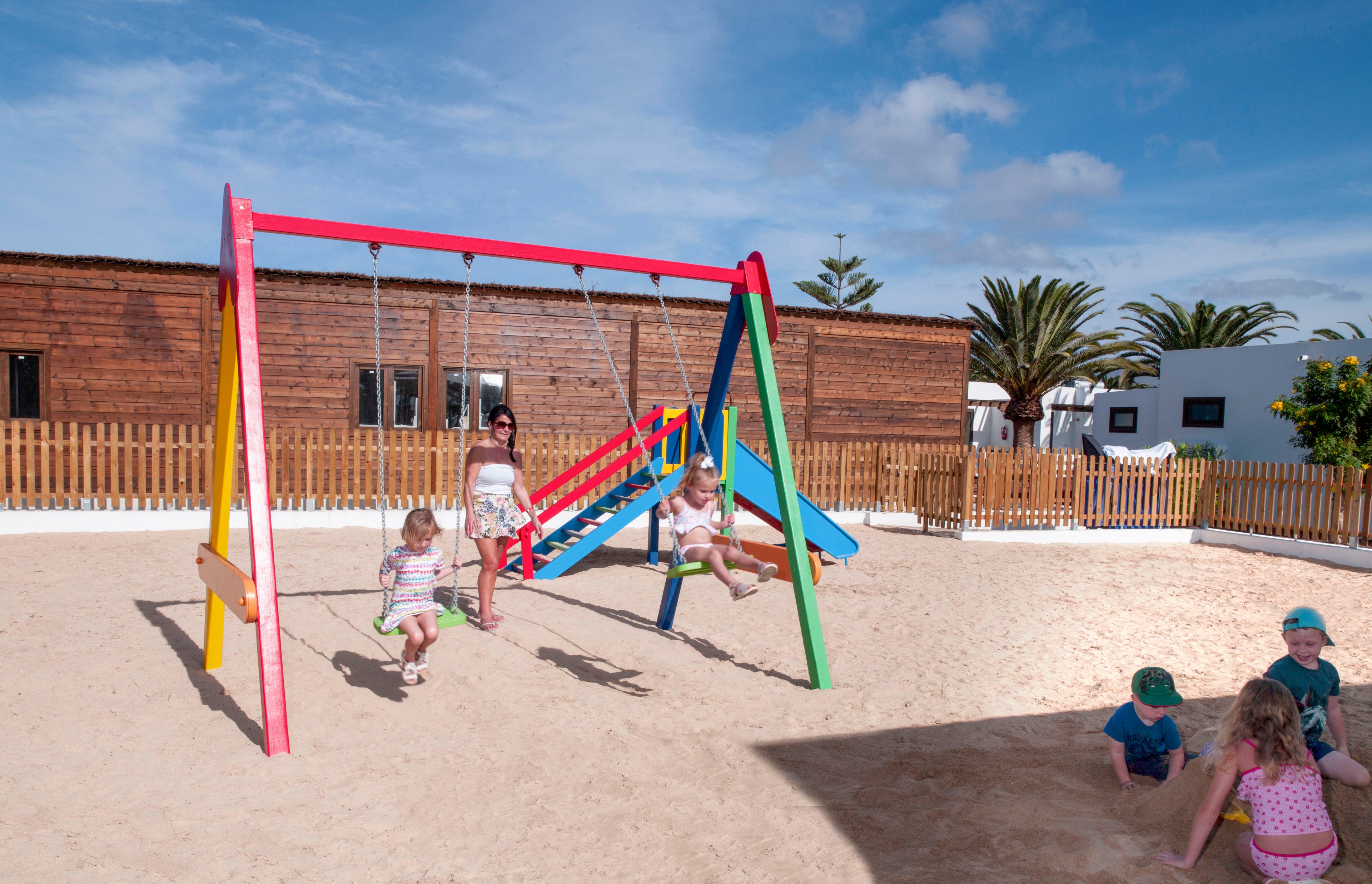 Several young children play together on a brightly colored swing set in a sandy outdoor playground. A woman stands nearby supervising as kids swing and climb under a clear blue sky. Wooden buildings and a fence surround the play area, with palm trees visible in the background. The scene suggests a relaxed, sunny holiday or resort environment focused on family fun.