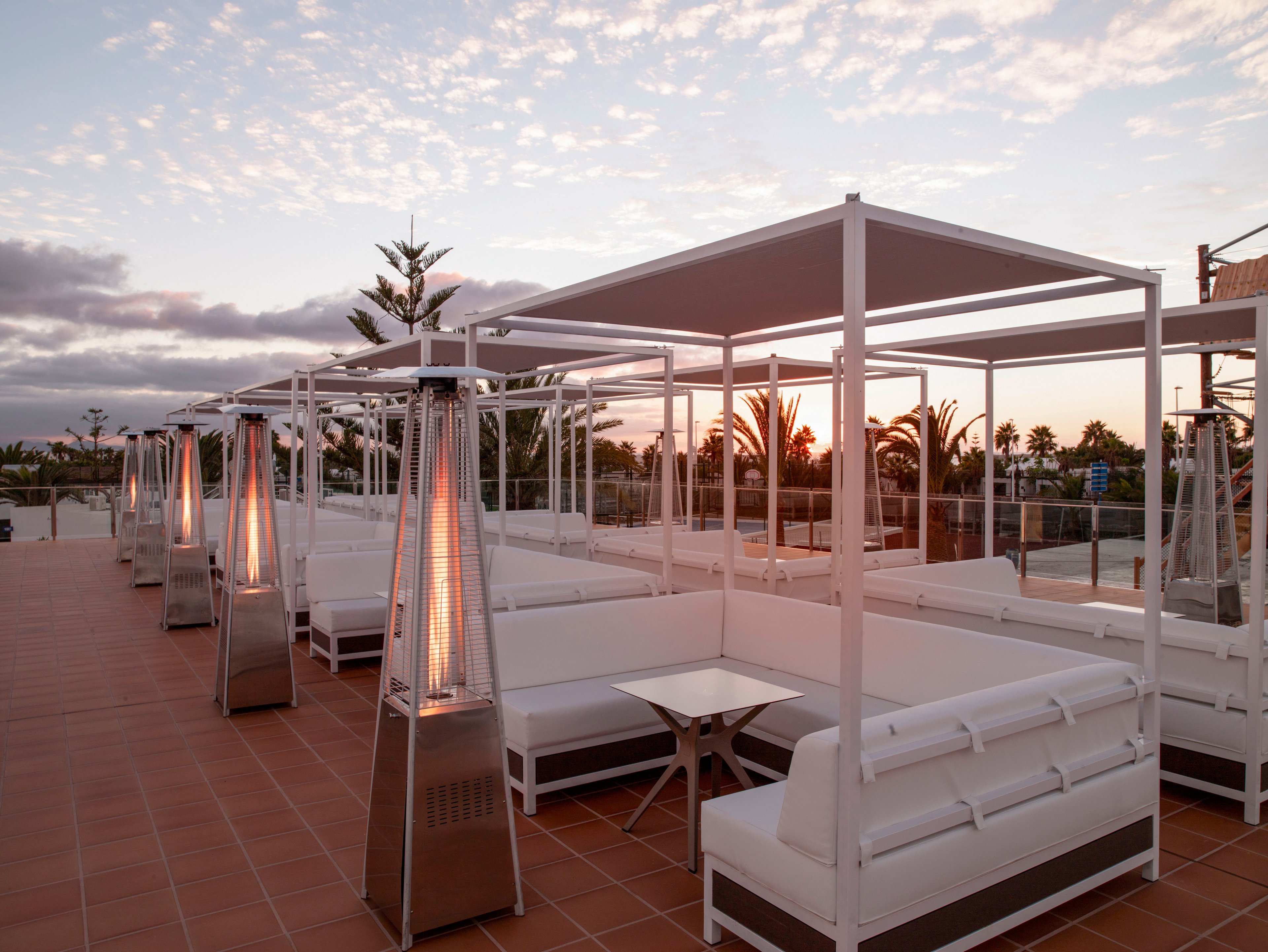 An elegant outdoor lounge area features white cabanas and cushioned seating arranged along a tiled terrace. Tall pyramid-style patio heaters glow warmly, creating a cozy atmosphere as the sun sets in the background. Palm trees and a soft, colorful sky add a resort-like feel to the scene. The space appears calm and inviting, ideal for evening relaxation or social gatherings.