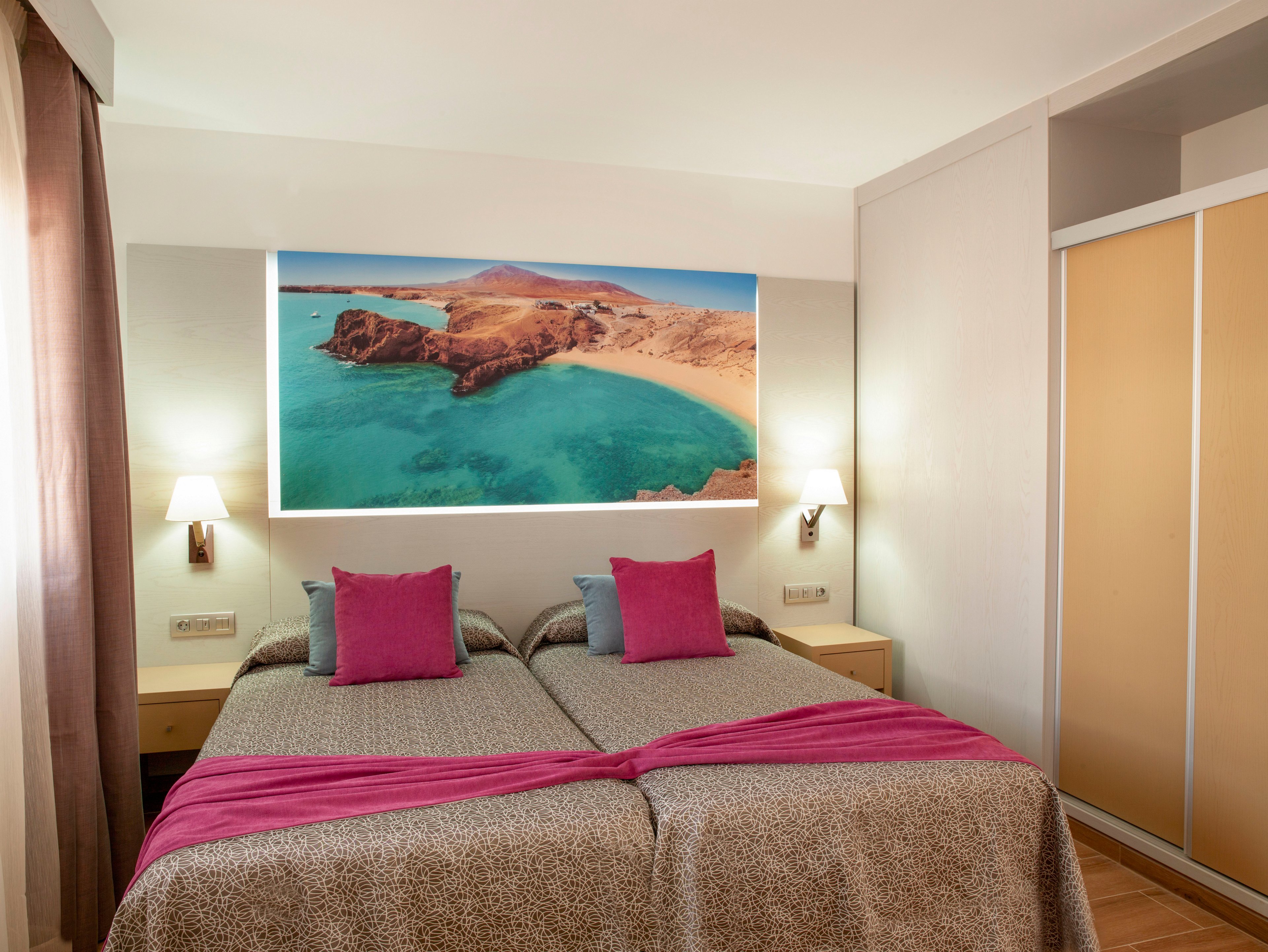 A cozy hotel room features two twin beds pushed together, topped with patterned bedspreads and bright pink cushions and runners. The space is warmly lit by wall-mounted lamps on either side of a large beach and ocean artwork above the headboard. Neutral walls, light wood bedside tables, and a sliding door wardrobe create a clean, modern look. Soft natural light filters in from a window with sheer curtains, enhancing the inviting atmosphere.