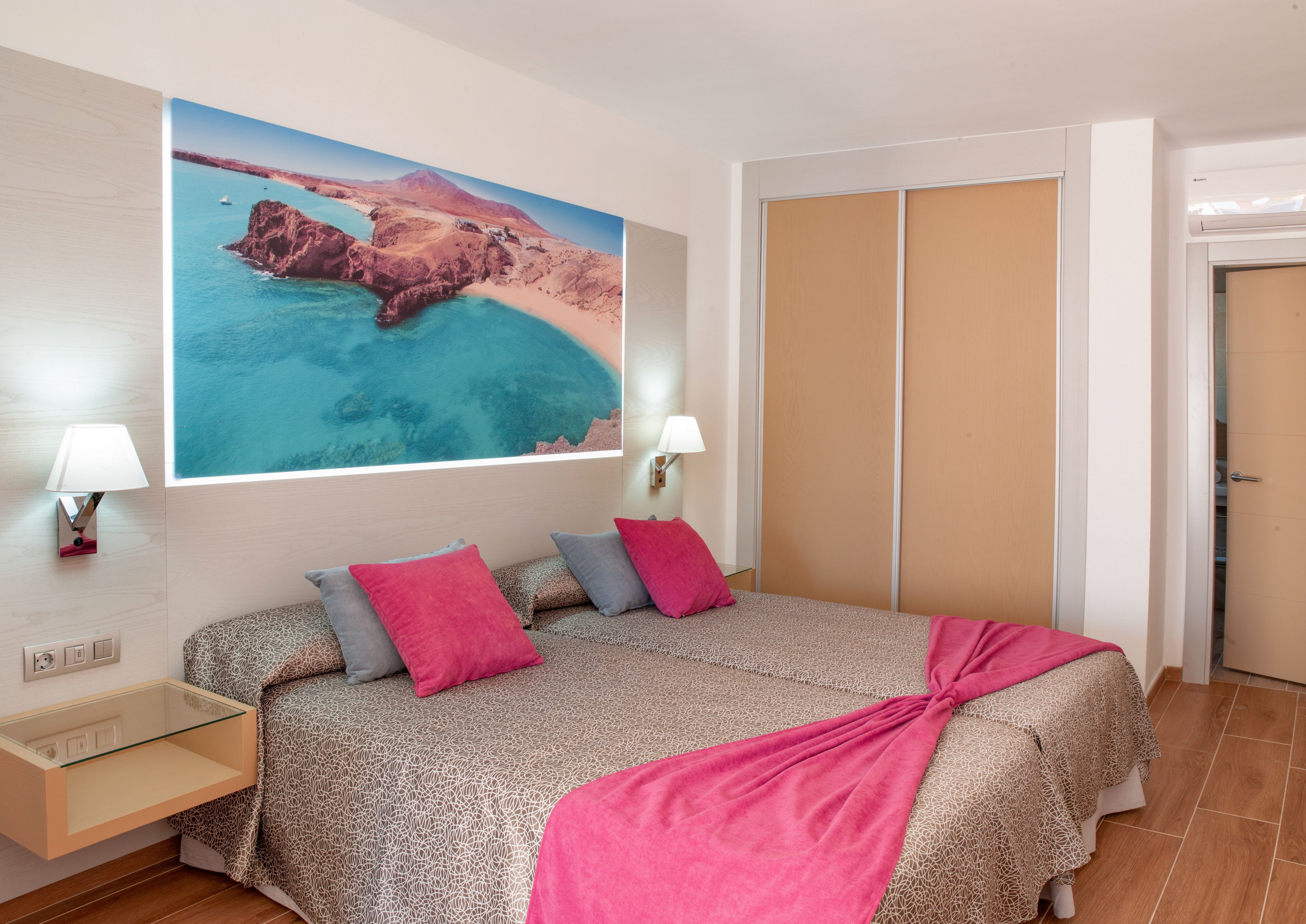 A contemporary hotel room features two twin beds pushed together, dressed in neutral patterned bedding with bright pink accents. Colorful throw pillows and a pink bed runner add contrast to the otherwise minimalist decor. A large aerial beach photograph hangs above the beds, creating a coastal theme. Wall-mounted bedside lamps and built-in shelves complete the clean, modern interior design.