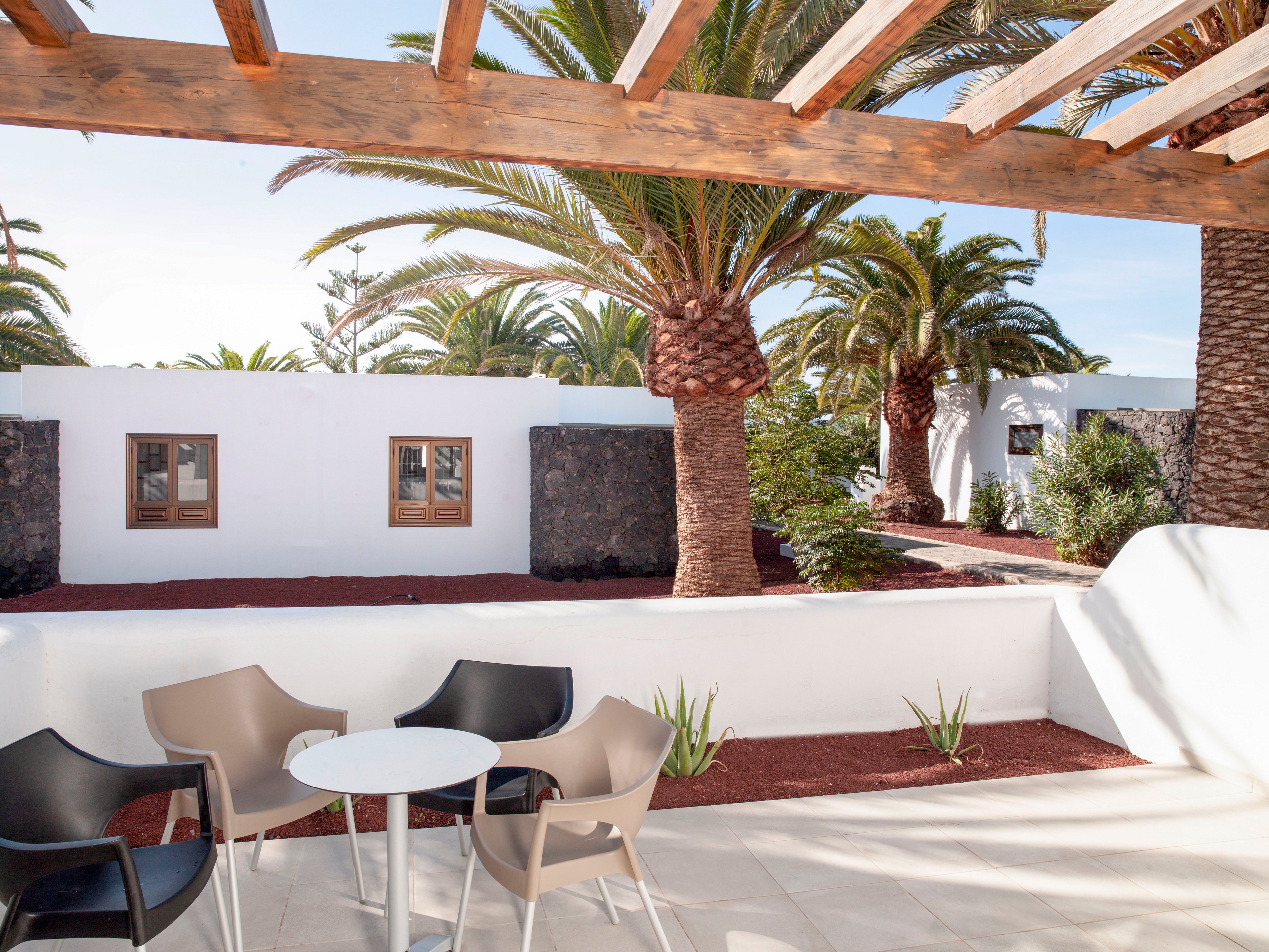 A bright outdoor terrace features modern patio chairs around a small round table. White walls and wooden pergola beams frame the space, creating a clean, minimalist look. Tall palm trees and volcanic stone accents add a tropical, resort-like atmosphere. The scene is sunlit and inviting, suggesting a relaxing holiday or leisure setting.