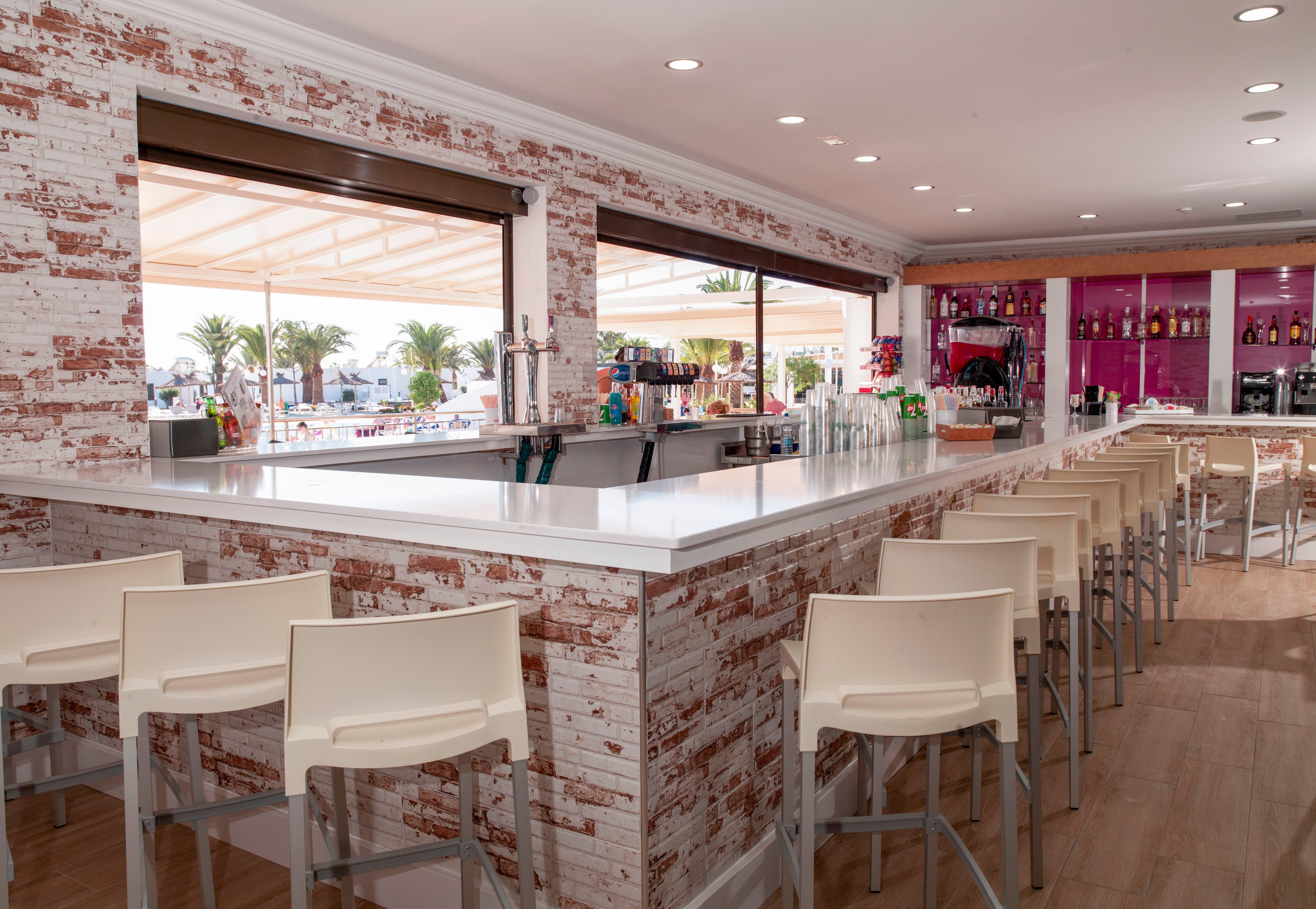 A contemporary bar interior features a large white counter surrounded by sleek white bar stools. The space has a rustic brick-style finish and light wood flooring, with colorful bottles displayed on shelves behind the bar. Large open windows reveal an outdoor terrace area with palm trees and bright daylight. The overall atmosphere is clean, inviting, and relaxed, suitable for socializing by the beach.