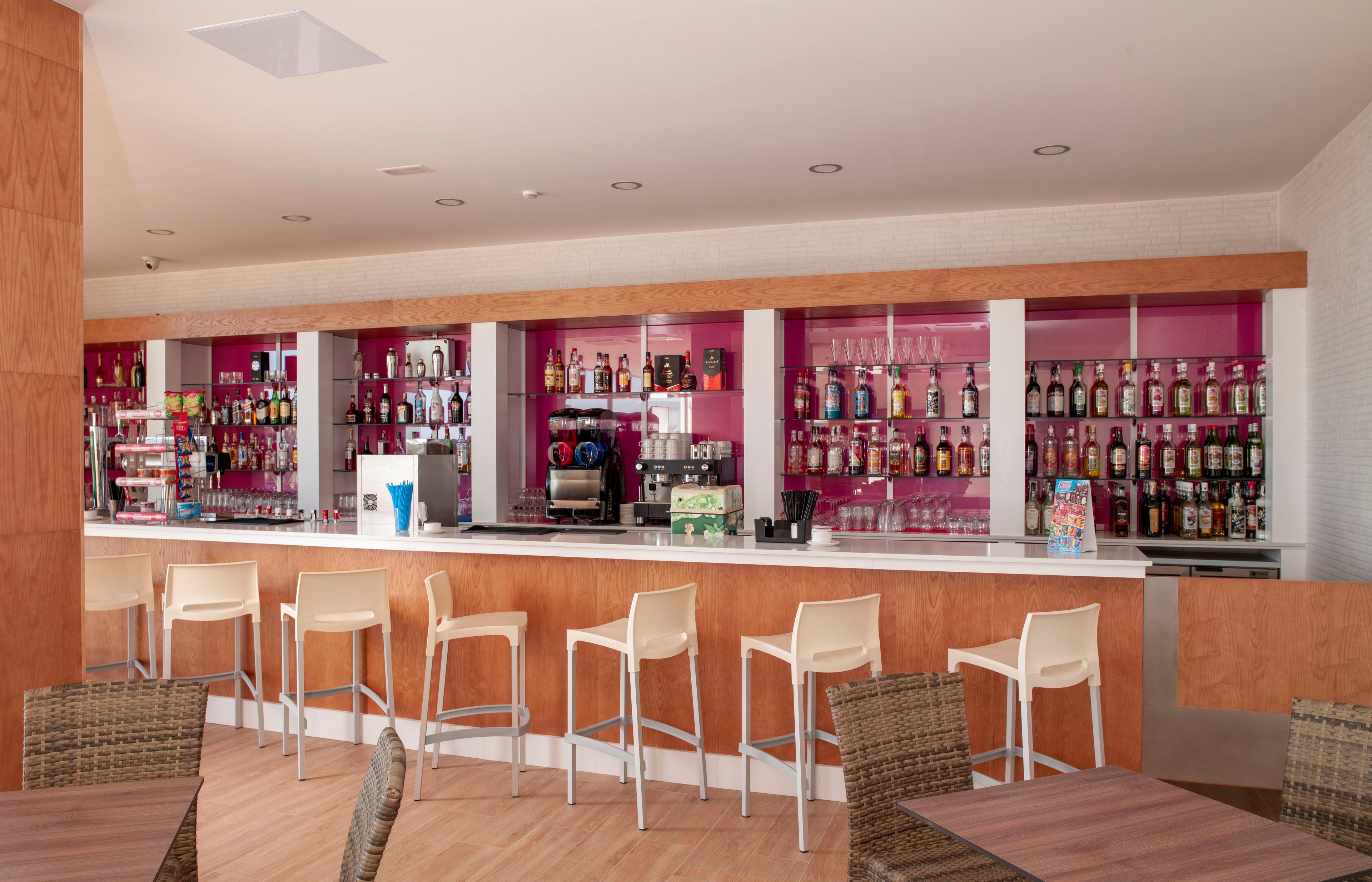 A contemporary bar interior features a long white counter lined with light-colored bar stools. Behind the counter, shelves filled with assorted liquor bottles and glassware are set against a bright pink backdrop. A coffee machine and other bar equipment are visible, suggesting service of both alcoholic drinks and coffee. The foreground shows wicker chairs and wooden tables, creating a relaxed lounge atmosphere in a clean, modern setting.