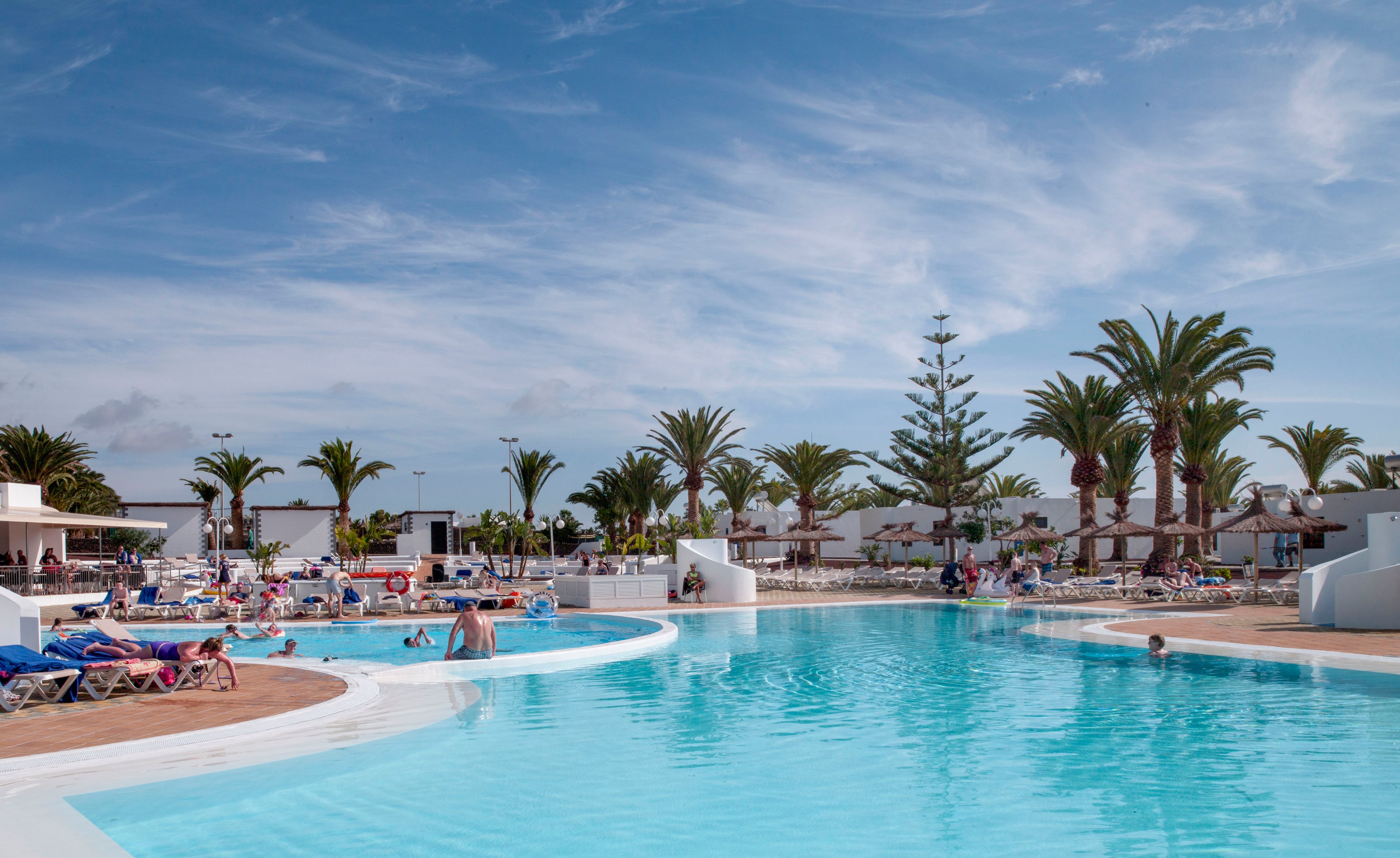 A large outdoor swimming pool is surrounded by sun loungers and people relaxing under a bright blue sky. Palm trees and low white resort buildings frame the scene, creating a tropical holiday atmosphere. Guests of various ages are sunbathing, swimming, and socializing around the pool area. The clear turquoise water and scattered umbrellas emphasize a warm, leisurely vacation setting.