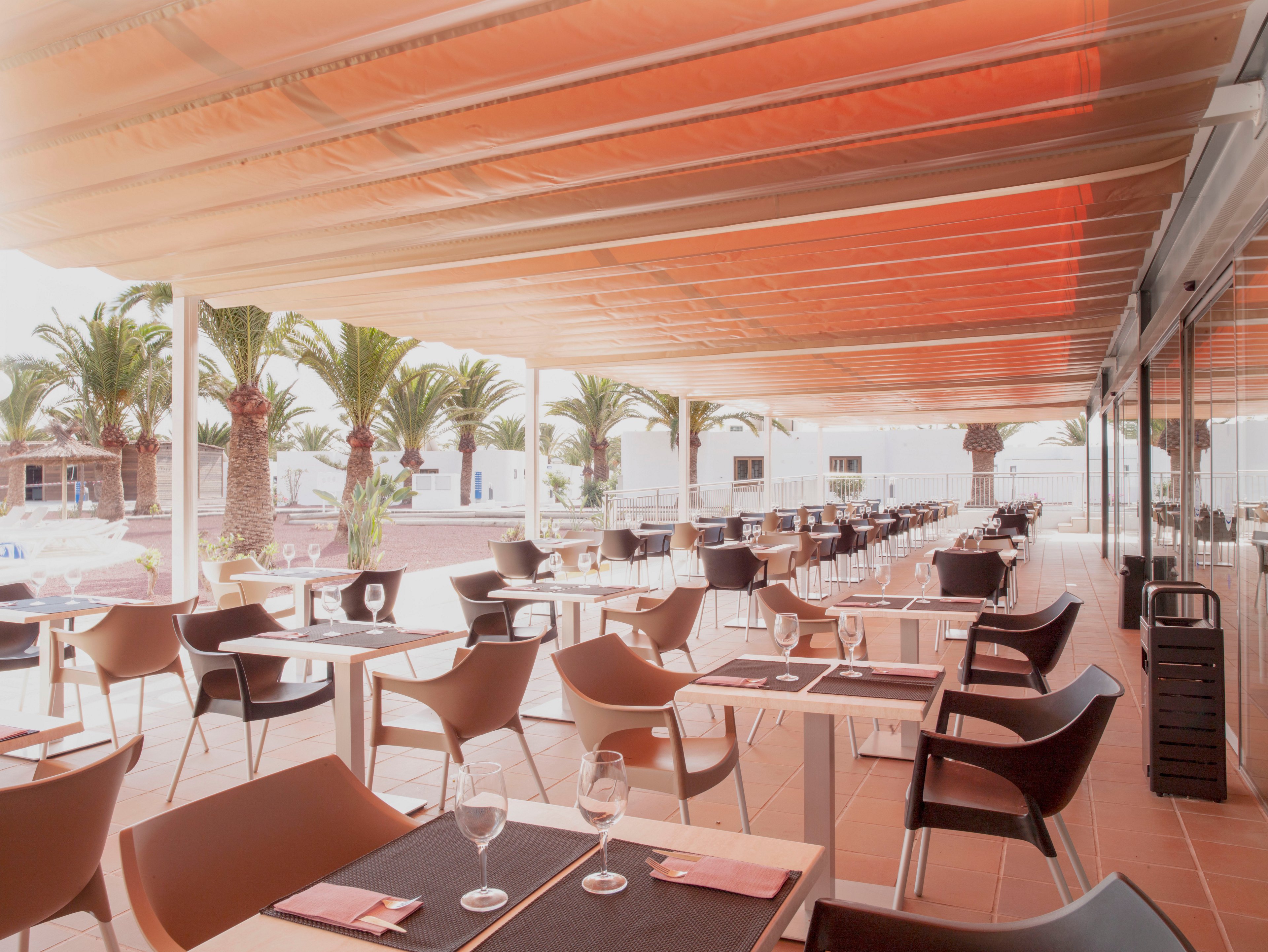 An open-air restaurant terrace features rows of modern tables and chairs neatly arranged under a large orange-tinted canopy. The setting appears calm and prepared for guests, with glassware and napkins placed on the tables. Palm trees line the background, suggesting a warm, resort-like environment. Reflections in the glass wall on the right add depth and emphasize the spaciousness of the dining area.