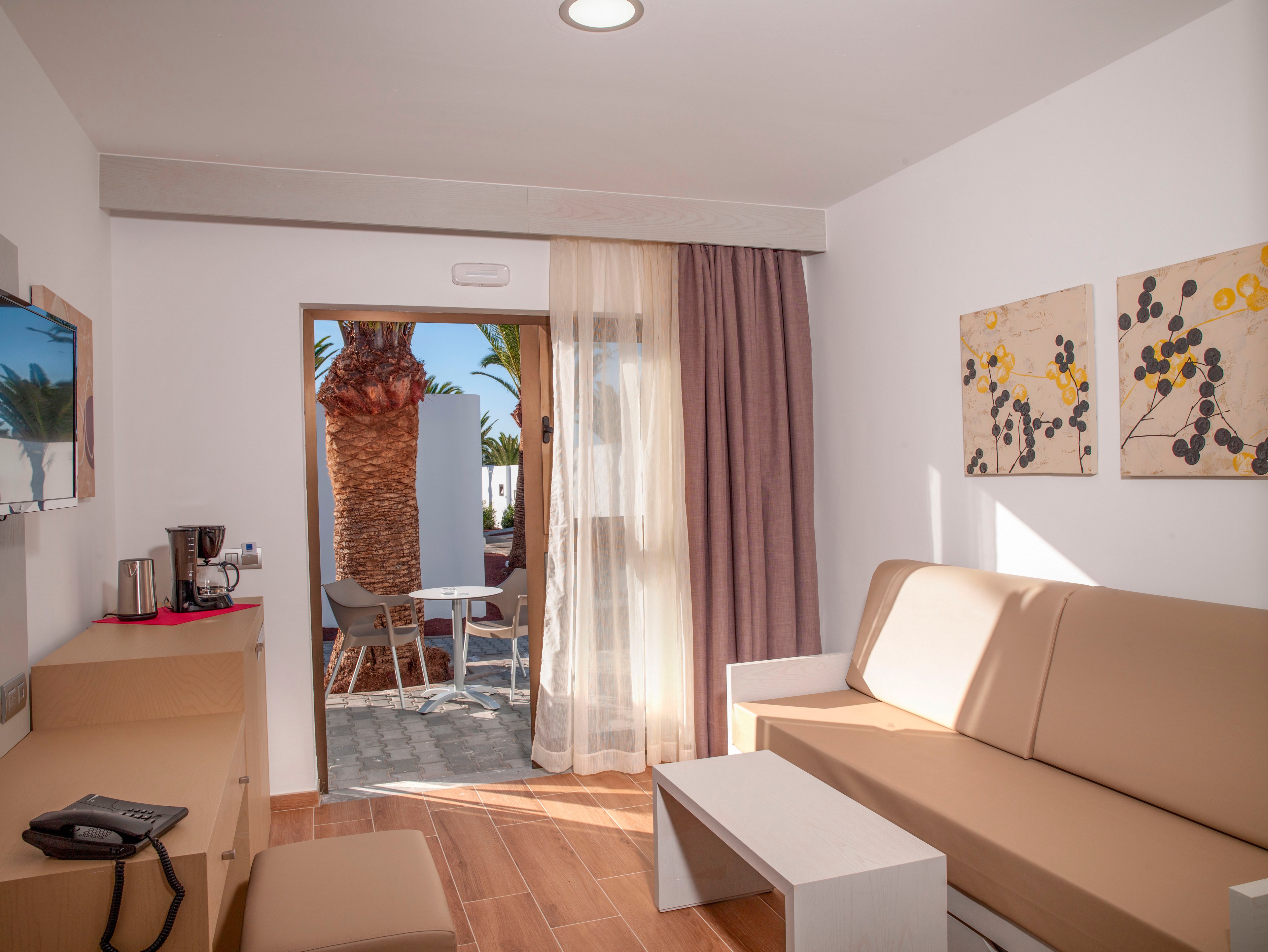 A contemporary hotel room features a beige sofa and minimalist furniture. Sunlight streams through sheer curtains and an open door leading to a terrace with outdoor chairs and palm trees. Warm wooden flooring and abstract wall art add a cozy, modern touch. A small desk area with a phone and coffee setup completes the comfortable vacation setting.
