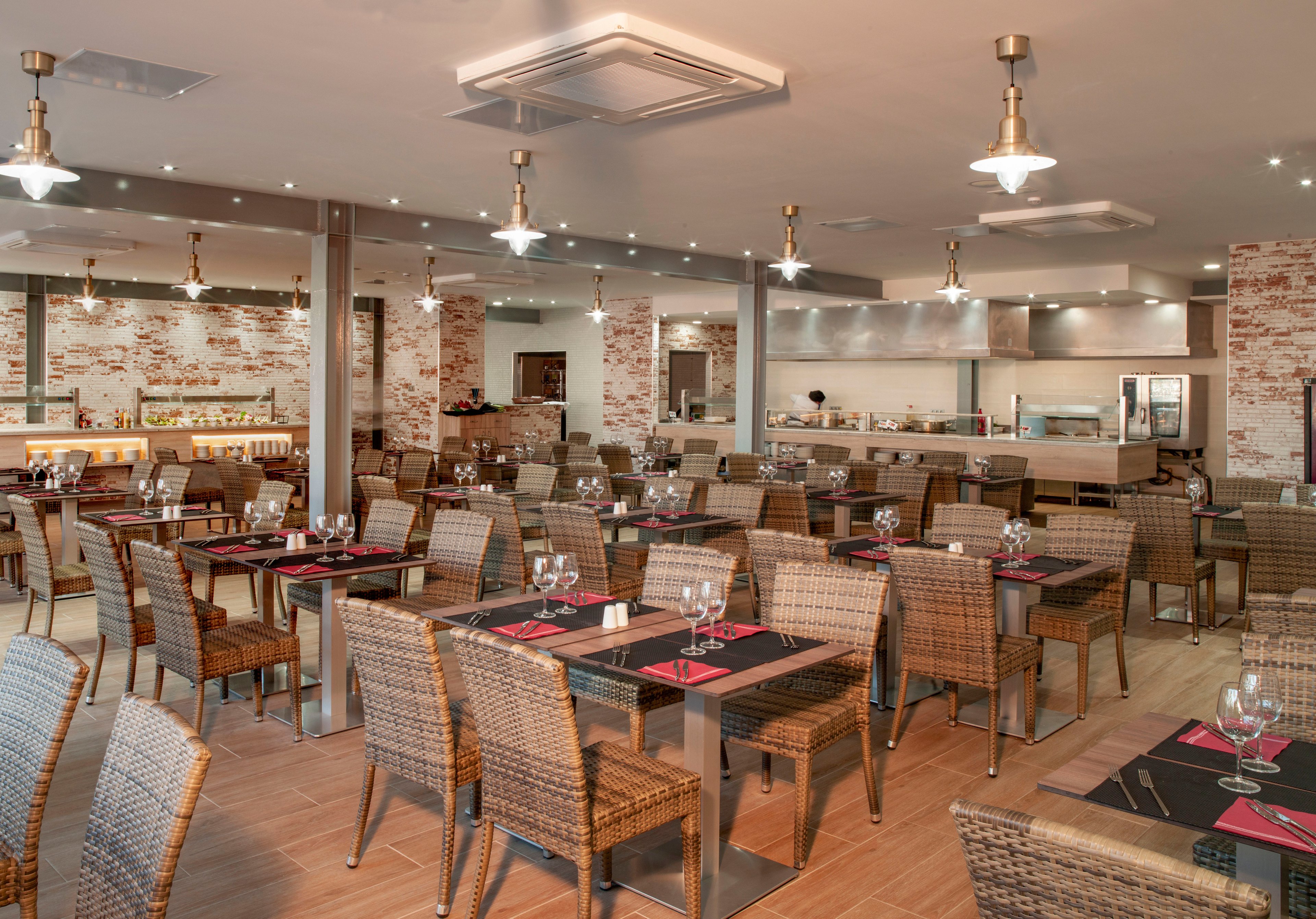 Spacious restaurant dining room with neatly arranged tables and wicker chairs. The setting features an open kitchen in the background with stainless steel equipment and warm brick accent walls. Tables are set with wine glasses, cutlery, and red placemats, suggesting readiness for guests. Ceiling lights and neutral tones create a contemporary, inviting atmosphere.