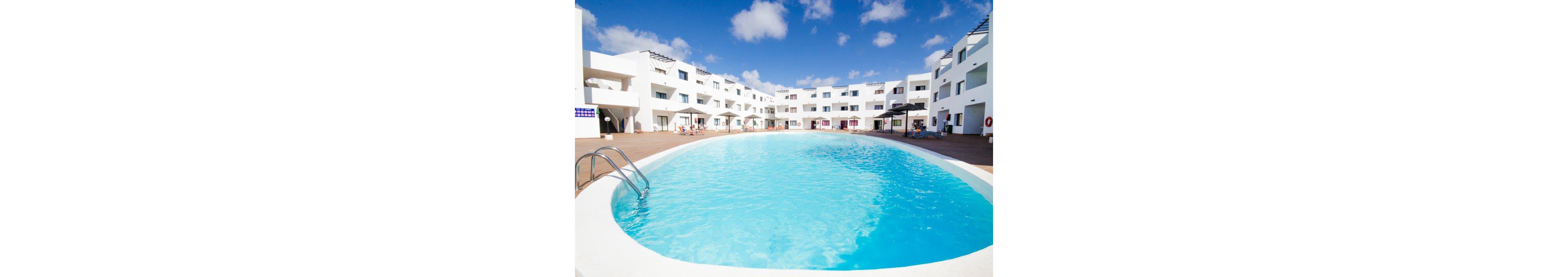 Large curved swimming pool sits at the center of a modern white apartment or hotel complex. Sun loungers and umbrellas line the spacious pool deck under a bright blue sky with scattered clouds. The water appears clear and inviting, reflecting the surrounding buildings. A metal ladder provides access to the pool, suggesting a relaxing vacation or resort setting.
