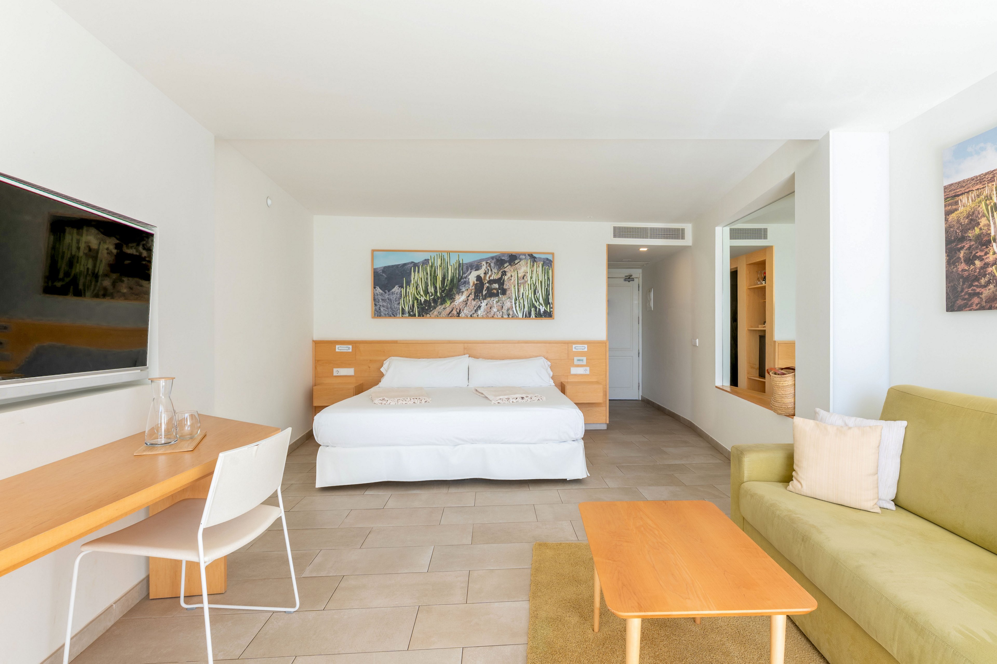 A spacious, modern hotel room features a large double bed centered against a wooden headboard. The room is decorated in light neutral tones with tiled flooring and minimal furnishings, including a green sofa, wooden coffee table, and a simple desk with a white chair. Wall art depicting nature scenes adds color to the otherwise clean, contemporary interior. Natural light brightens the space, creating an inviting and comfortable atmosphere.