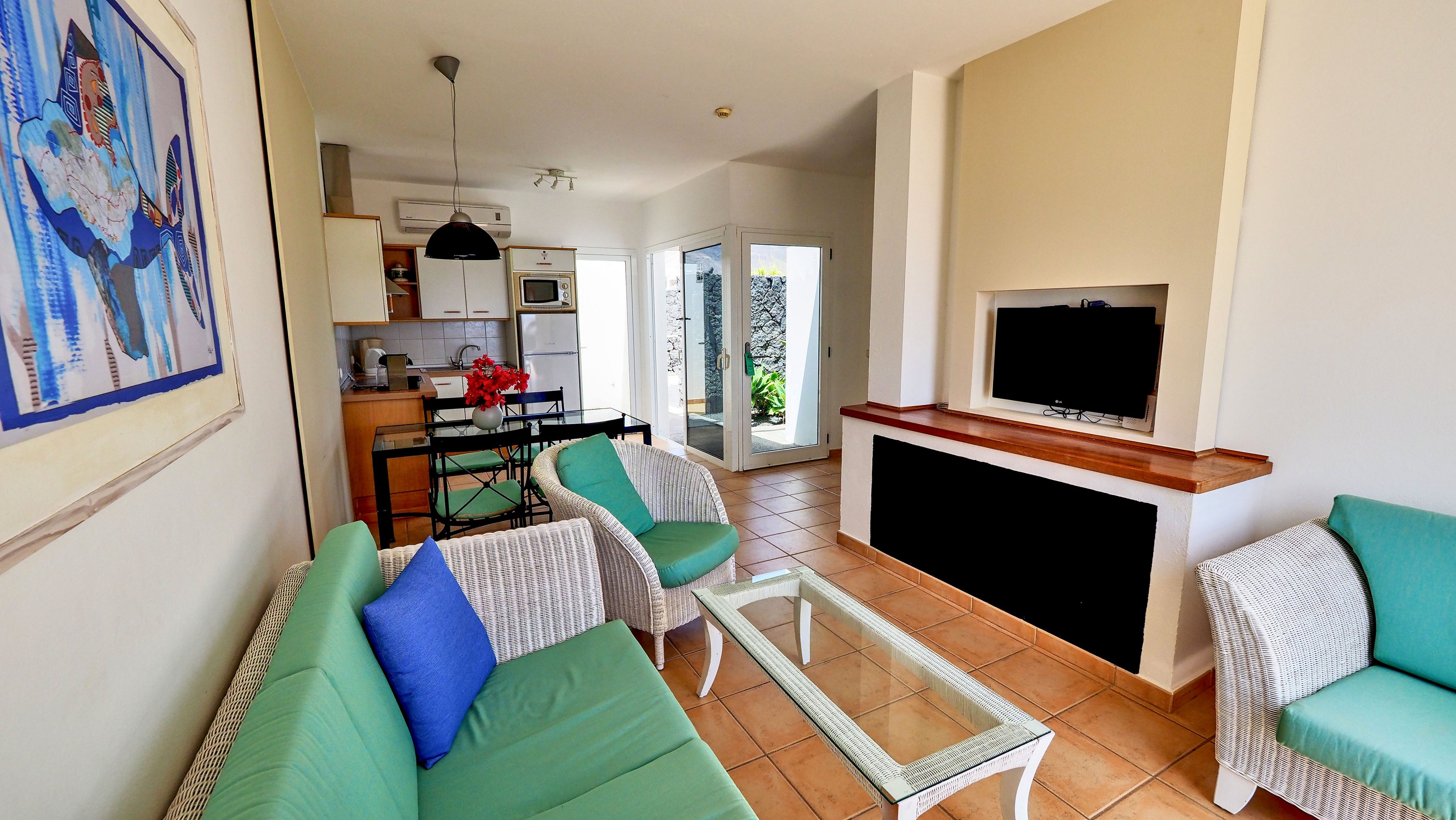 A sunlit living room features wicker seating with green cushions and a glass coffee table on tiled floors. A flat-screen television is mounted in a recessed wall opposite the sofa. In the background, an open-plan kitchen and dining area include white cabinets, appliances, and a small table with chairs. Large glass doors lead to an outdoor space, adding natural light to the airy interior.