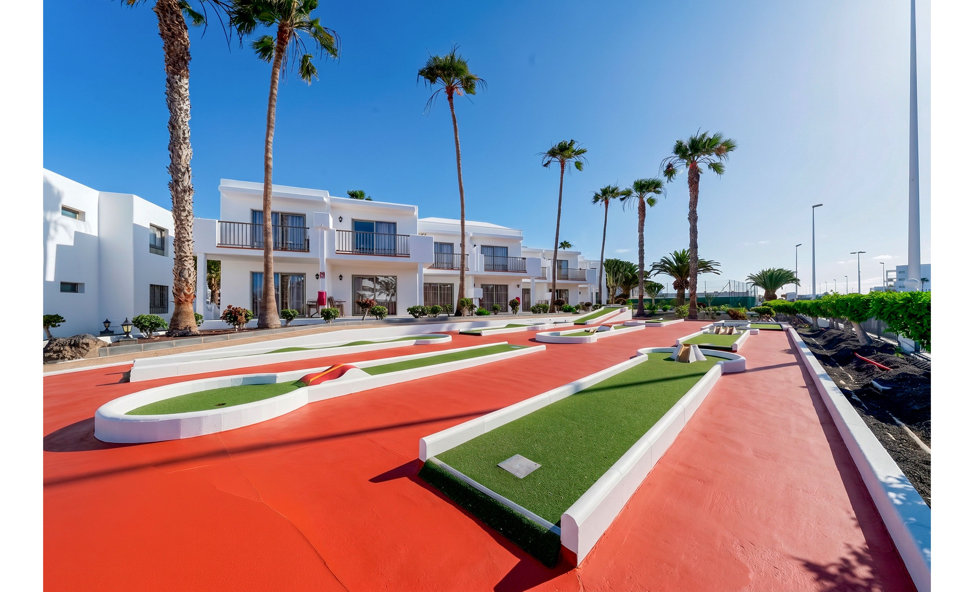 A brightly colored mini golf course is set in front of modern white resort buildings under a clear blue sky. Tall palm trees line the walkway, adding a tropical atmosphere to the scene. The putting lanes feature green artificial turf bordered by white edges on a vivid red surface. The overall setting appears clean, sunny, and inviting, suggesting a vacation or leisure destination.