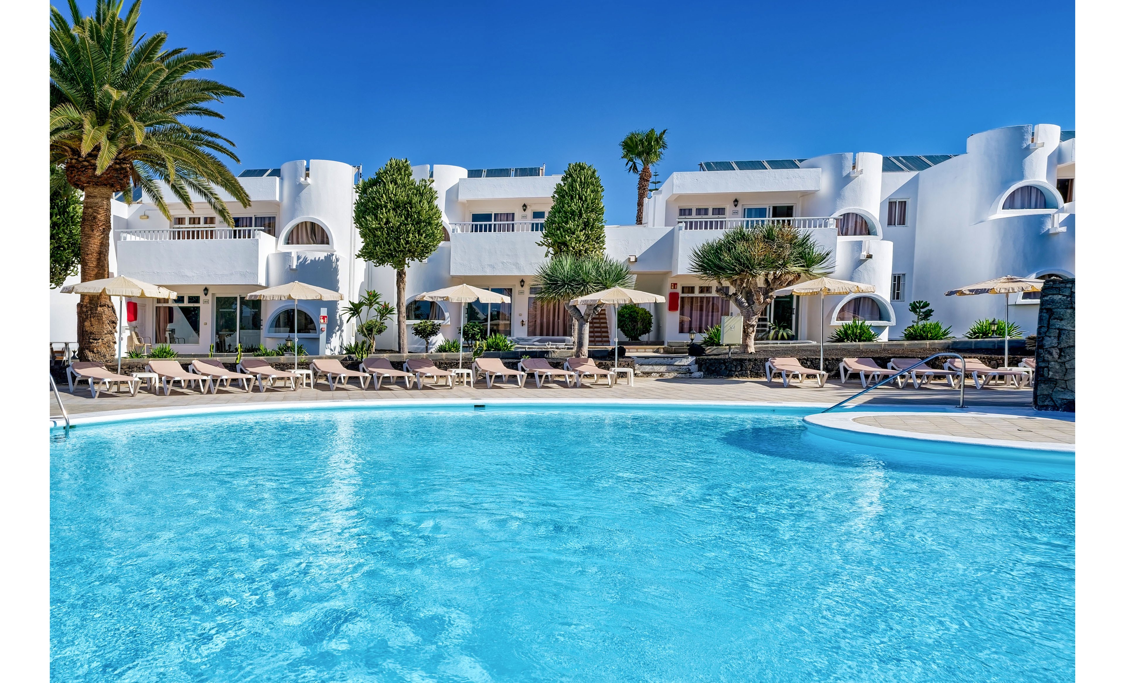 A bright blue swimming pool sits in front of a white, Mediterranean-style resort building. Numerous sun loungers and beige umbrellas line the poolside terrace, suggesting a relaxing holiday setting. Palm trees and manicured greenery add a tropical feel to the scene. The clear sky and vivid colors create a warm, inviting atmosphere ideal for vacation imagery.