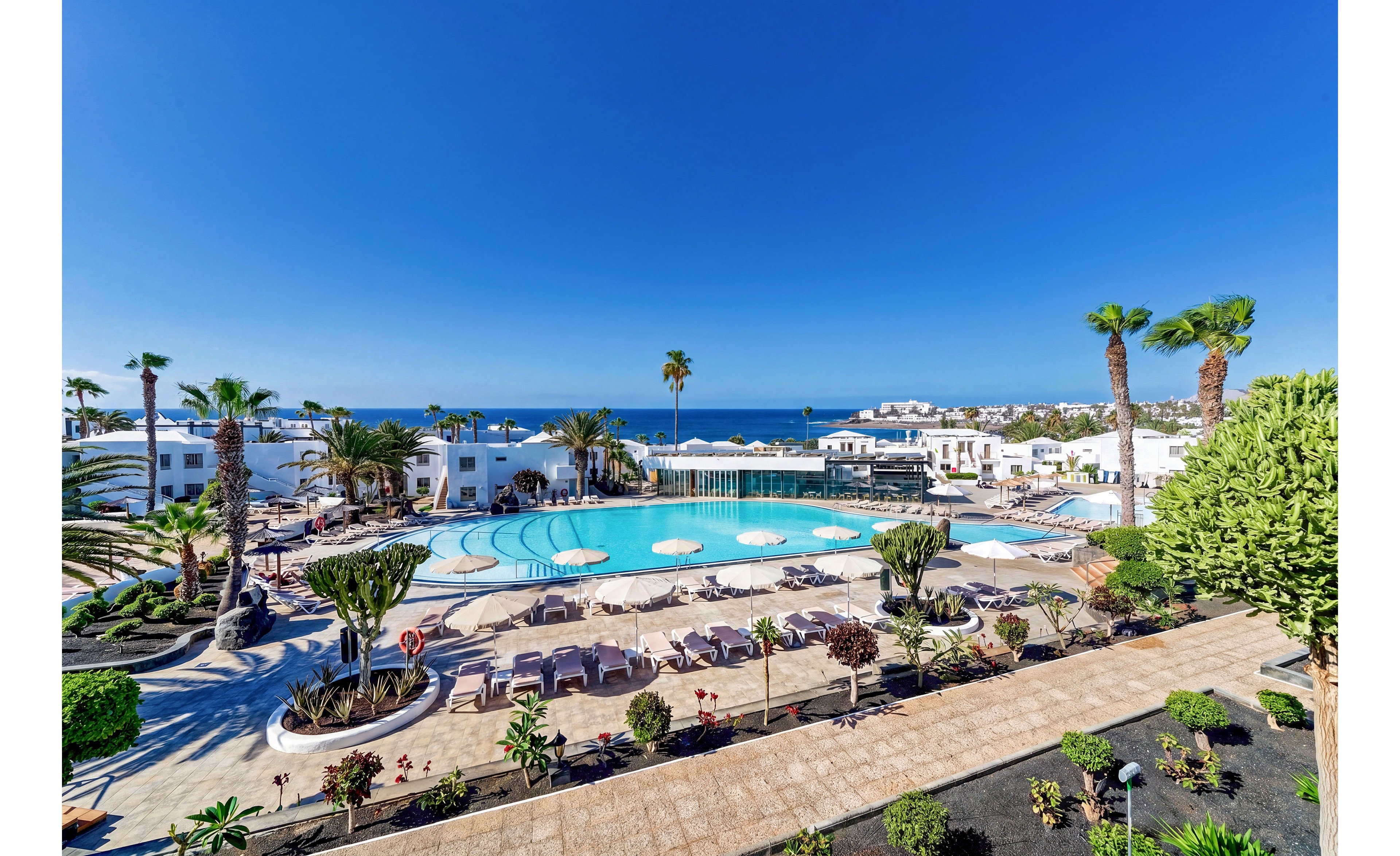 A bright seaside resort features a large circular swimming pool surrounded by sun loungers and umbrellas. White low-rise buildings frame the pool area, with palm trees and manicured gardens adding lush greenery. The scene overlooks a deep blue ocean under a clear cloudless sky, suggesting a warm vacation destination. Pathways and landscaped beds in the foreground lead the eye toward the pool and distant coastline.