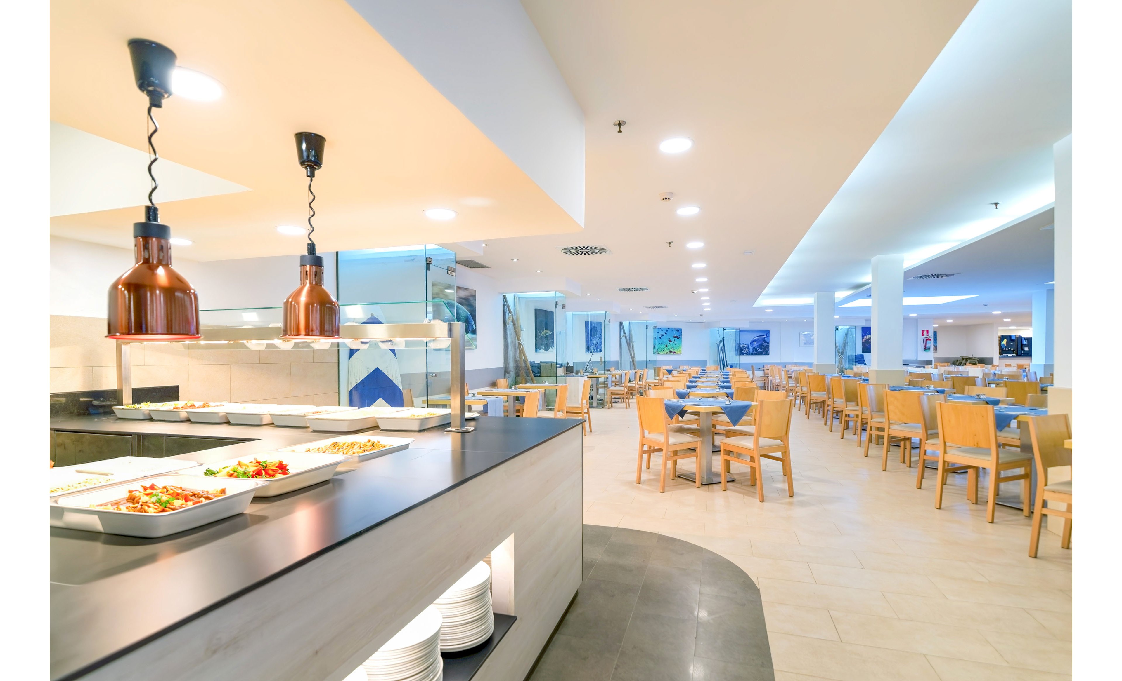 A spacious self-service cafeteria features a sleek buffet counter with trays of fresh food. Warm pendant lights hang above the serving area, contrasting with the bright white ceiling and walls. Rows of light wooden tables and chairs extend into the background, creating an orderly dining space. The overall atmosphere is clean, modern, and welcoming, suitable for large groups or casual meals.