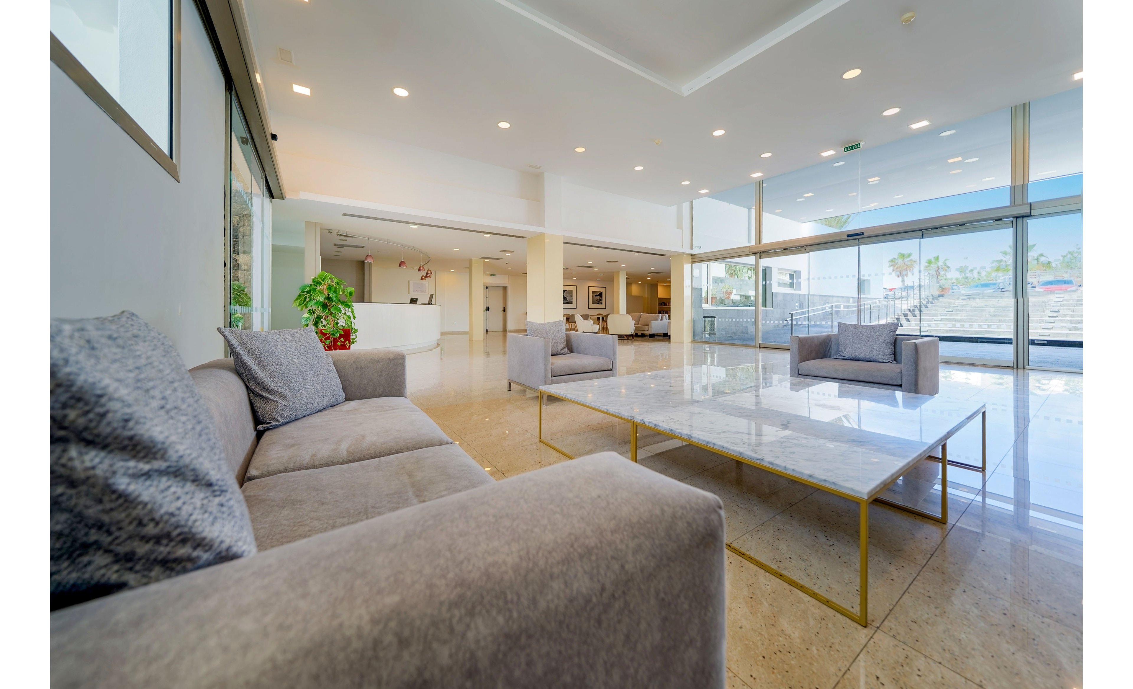 Spacious contemporary lobby interior featuring gray upholstered sofas around a large low marble table with slim metallic legs. The room has polished stone flooring, white walls, and recessed ceiling lighting that creates a clean, minimalist atmosphere. Large floor-to-ceiling windows on one side let in abundant natural light and offer views of an outdoor area. A potted plant and framed artwork add subtle decorative accents to the otherwise sleek, modern space.