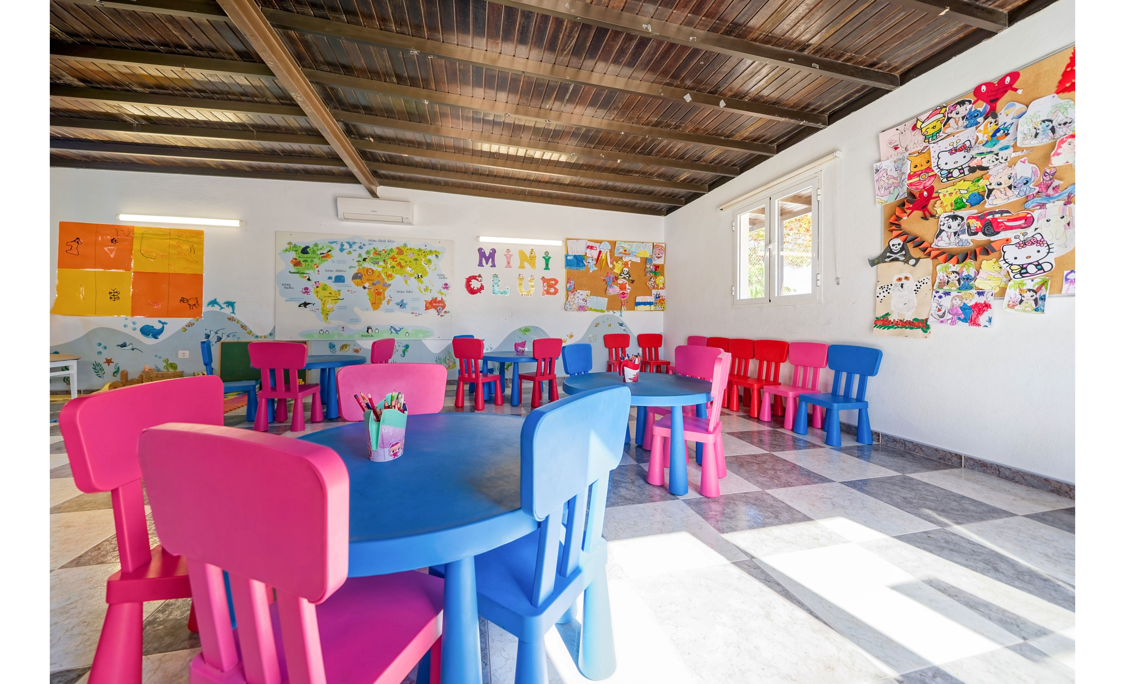 A cheerful children’s playroom is furnished with bright pink and blue plastic tables and chairs arranged around the room. The walls are decorated with world maps, educational posters, and a large collage of cartoon drawings. Sunlight streams in through the windows, illuminating the tiled floor and the art supplies placed on the tables. The space feels inviting and creative, designed for kids’ activities and learning.