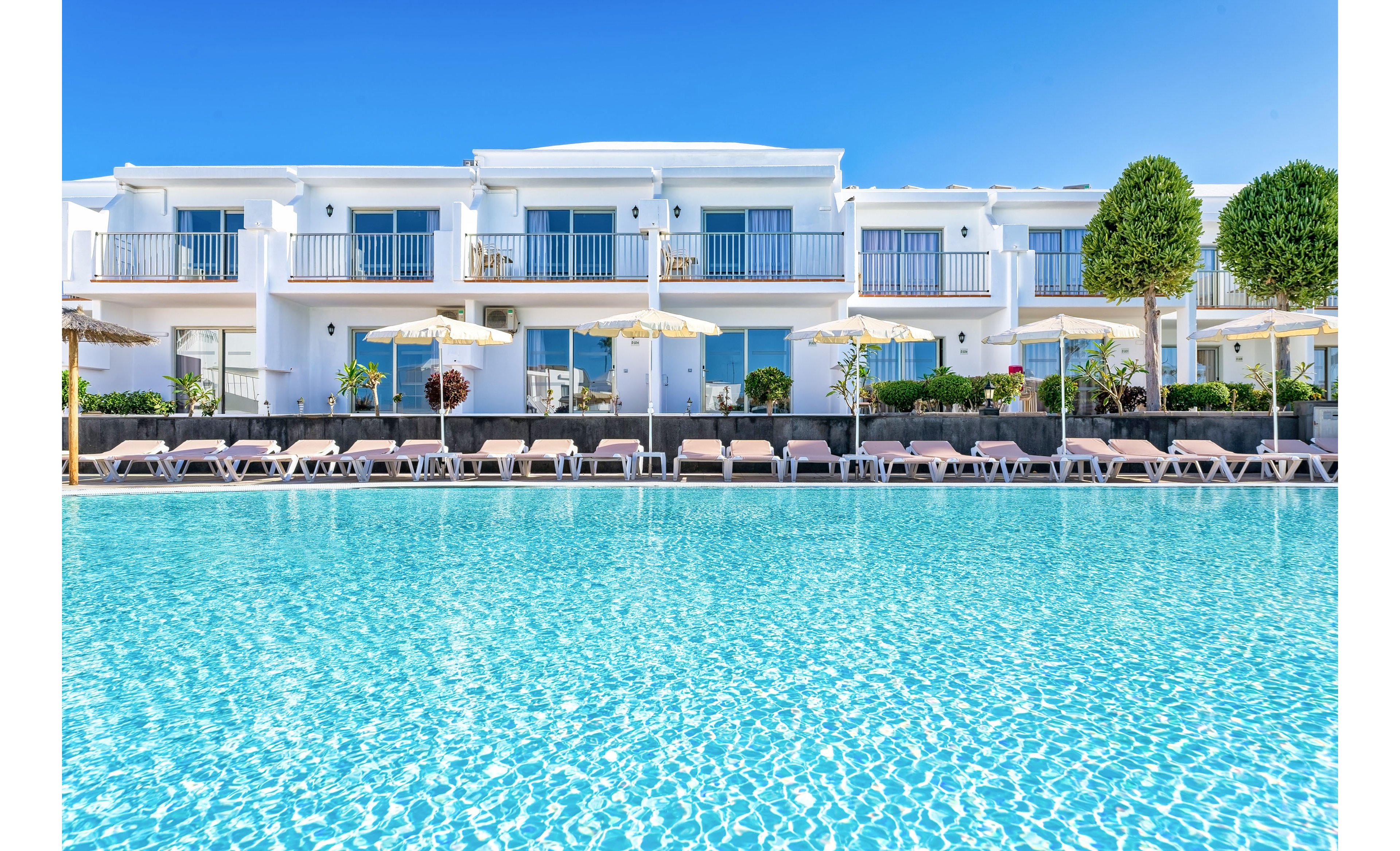 A bright, modern resort hotel features a large turquoise outdoor swimming pool in the foreground. White two-story buildings with balconies and glass doors line the poolside, creating a clean and contemporary look. Numerous sun loungers and parasols are arranged along the edge of the pool, suggesting a relaxing vacation setting. Clear blue sky and neatly trimmed trees enhance the sunny, tranquil atmosphere.