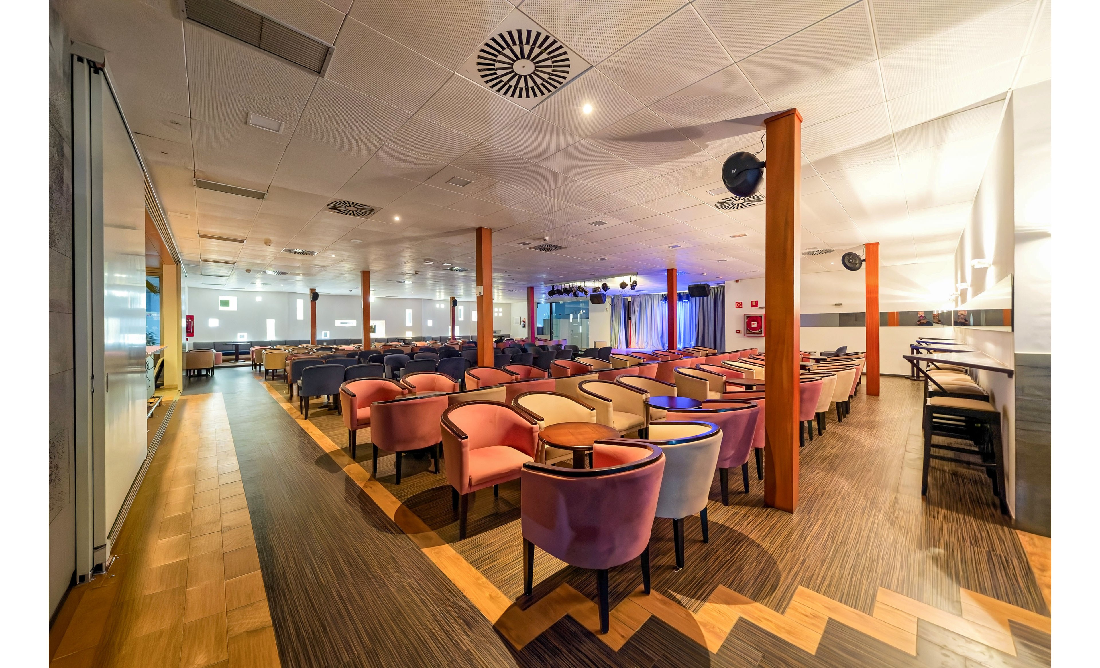Spacious indoor lounge set up for a conference or presentation with rows of colorful armchairs and small round tables. The room features a modern ceiling with recessed lighting and patterned ventilation panels, supported by wooden columns. A stage area with curtains and soft colored lights is visible at the far end, suggesting a venue for talks or performances. The floor combines wood and dark patterned surfaces, creating a contemporary, welcoming atmosphere.