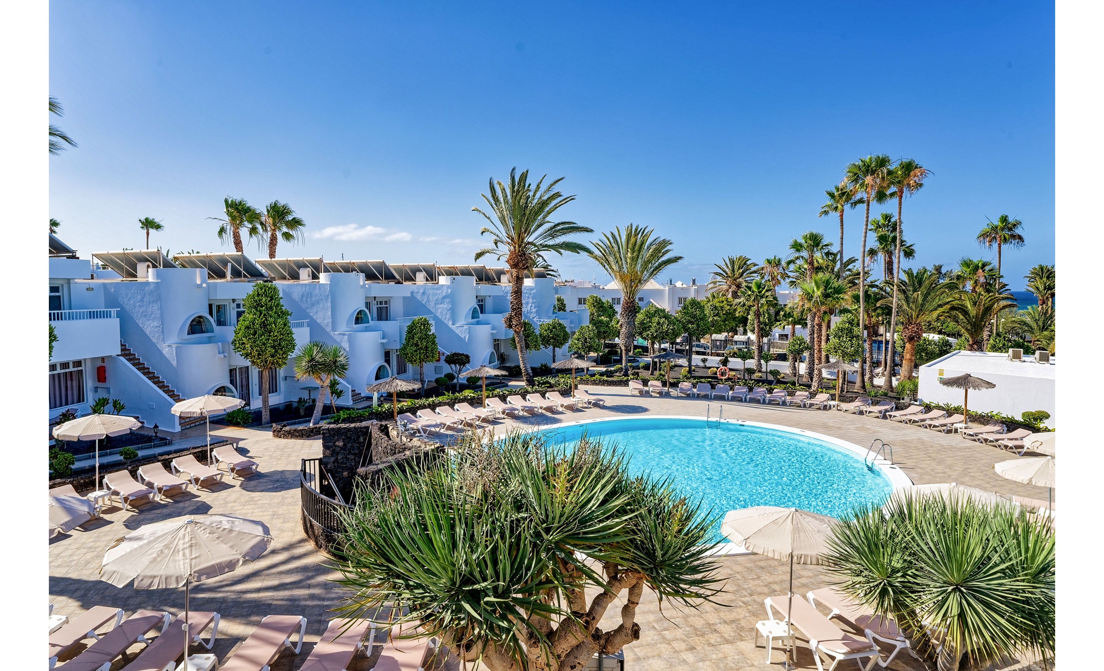 A circular outdoor swimming pool is surrounded by sun loungers and parasols in a bright resort setting. White villa-style buildings with terraces and balconies frame the pool area. Tall palm trees and lush greenery add a tropical atmosphere under a clear blue sky. The scene suggests a relaxing holiday destination with ample seating and sunny weather.