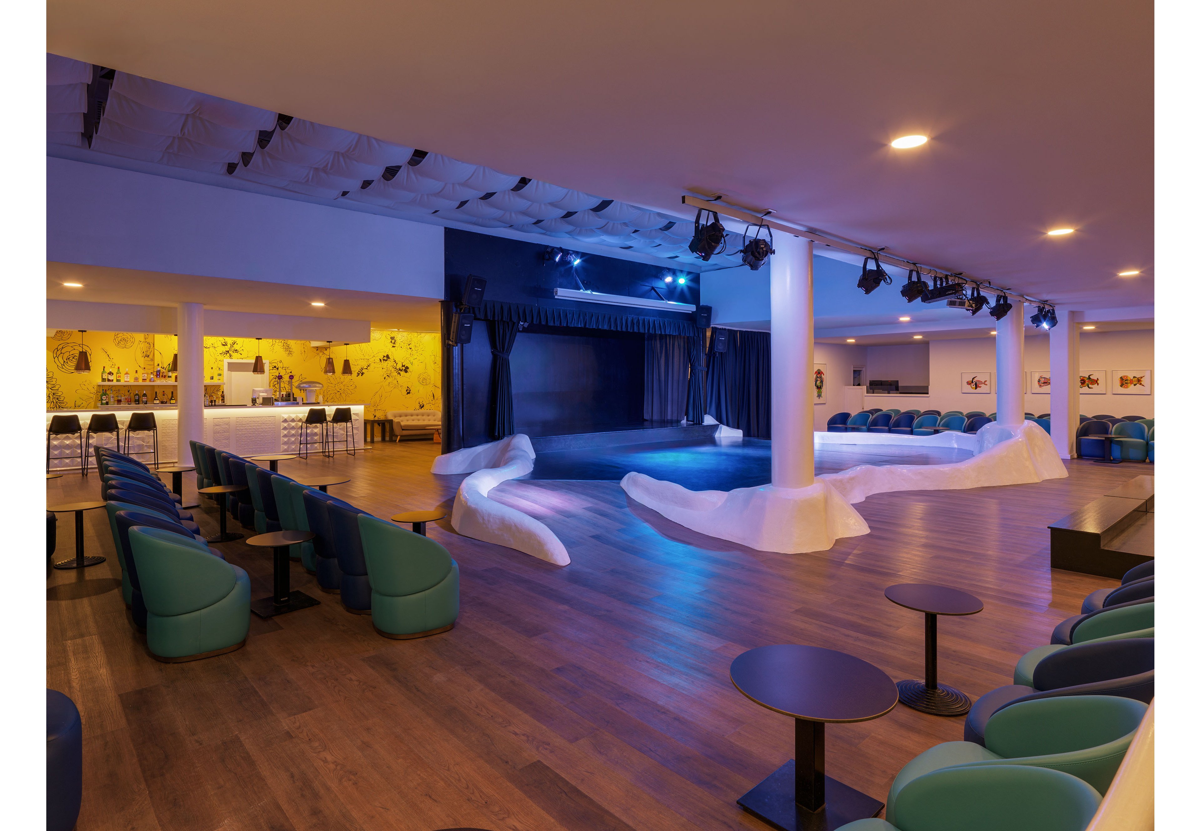 A contemporary indoor lounge theater features a central stage framed by sculptural white seating elements. Rows of teal armchairs and small round tables are arranged on a wooden floor, facing the stage. Warm ceiling lights contrast with cool blue stage lighting, creating a relaxed yet vibrant atmosphere. In the background, a bar or refreshment area is visible, adding to the social setting.