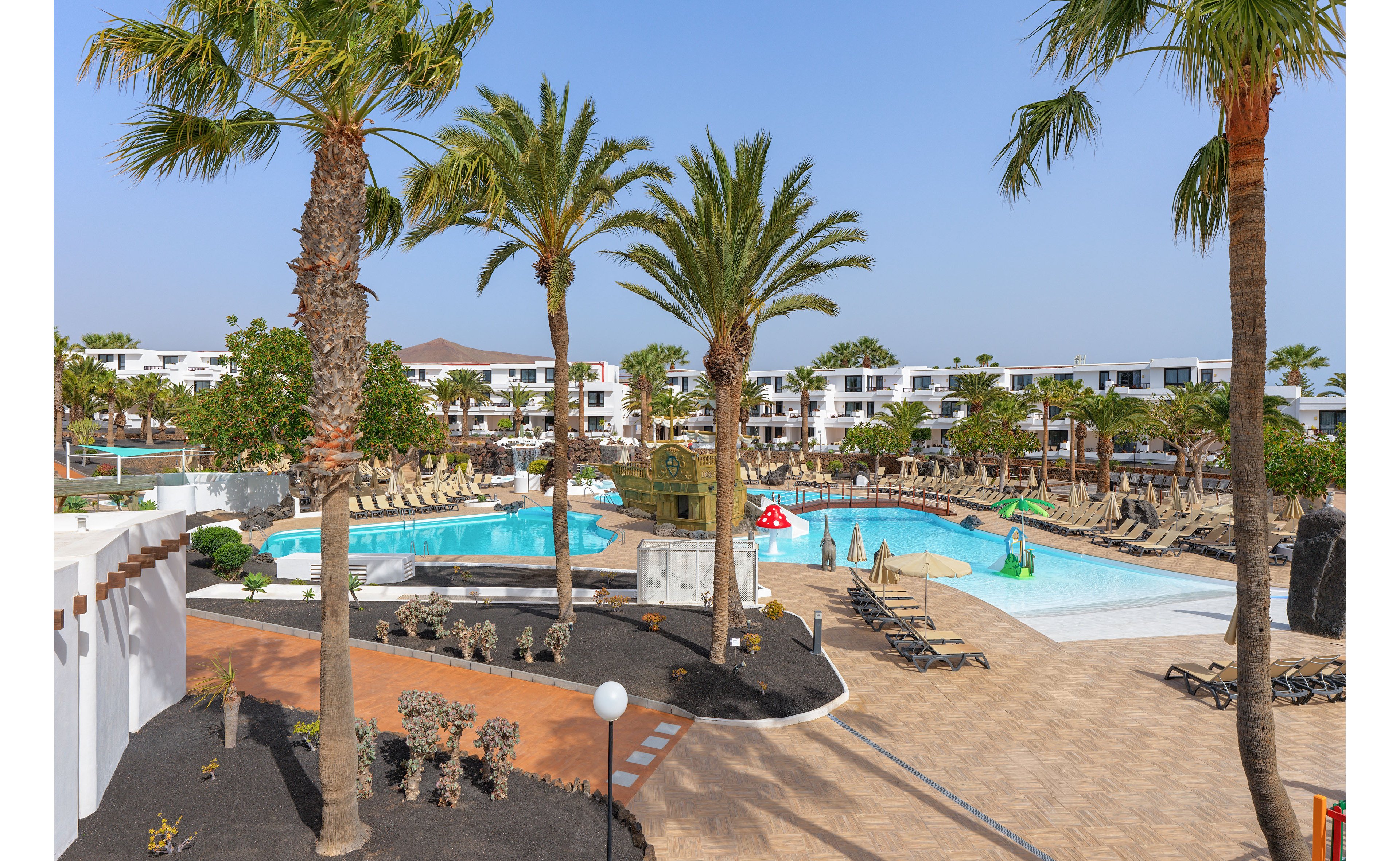 A large resort swimming pool is surrounded by tall palm trees and sun loungers under a clear blue sky. White low-rise buildings form a backdrop, giving the scene a modern vacation complex feel. The pool area includes a small water play structure and shallow sections suitable for families. Pathways, landscaped beds, and neatly arranged deck chairs emphasize a relaxed holiday atmosphere.