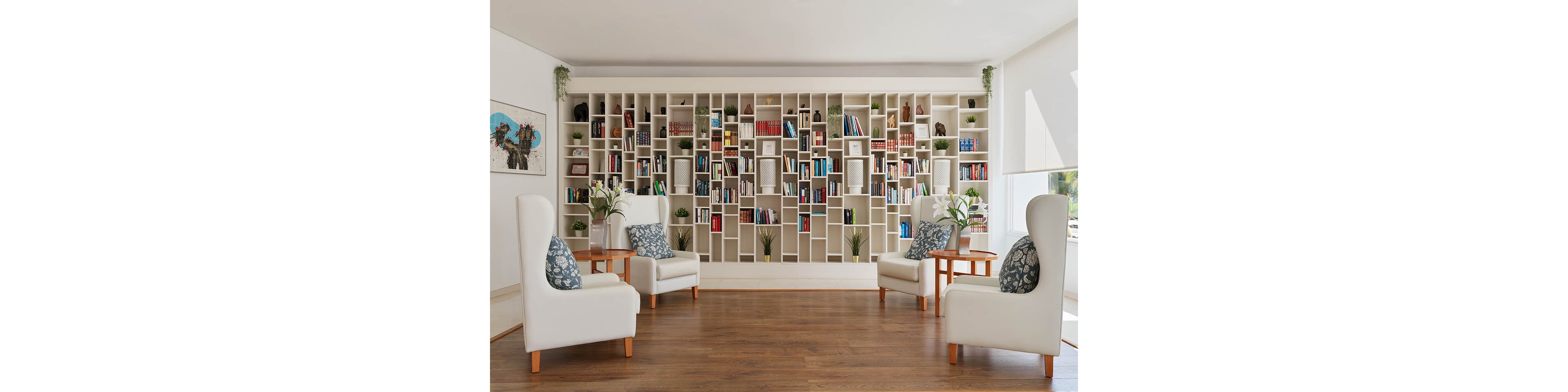 A contemporary reading lounge features a full wall of white bookshelves filled with books and decorative plants. Four white armchairs with patterned cushions are arranged in a conversational layout around small wooden side tables. The room has warm wooden flooring and bright natural light from large windows. Minimalist artwork and greenery add a calm, inviting atmosphere suitable for relaxing or reading.