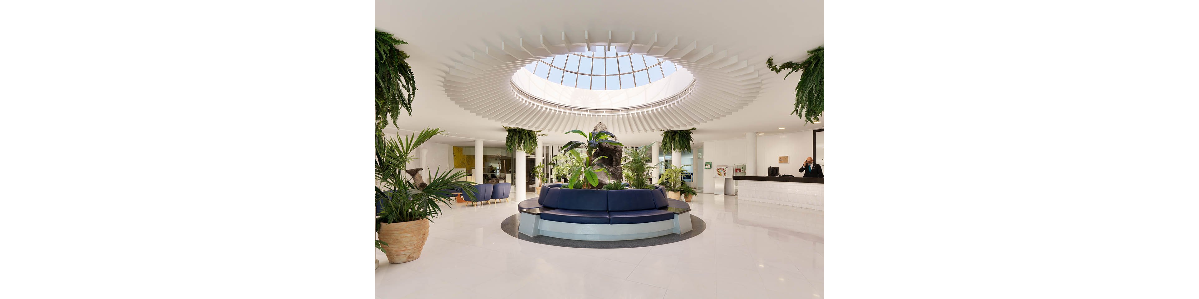 A spacious, contemporary hotel lobby features a large circular skylight above a central seating area. Blue curved sofas surround lush indoor plants, creating a welcoming focal point. The bright white floor and walls enhance the airy, minimalist design. A reception desk with staff is visible in the background, adding a sense of hospitality and service.