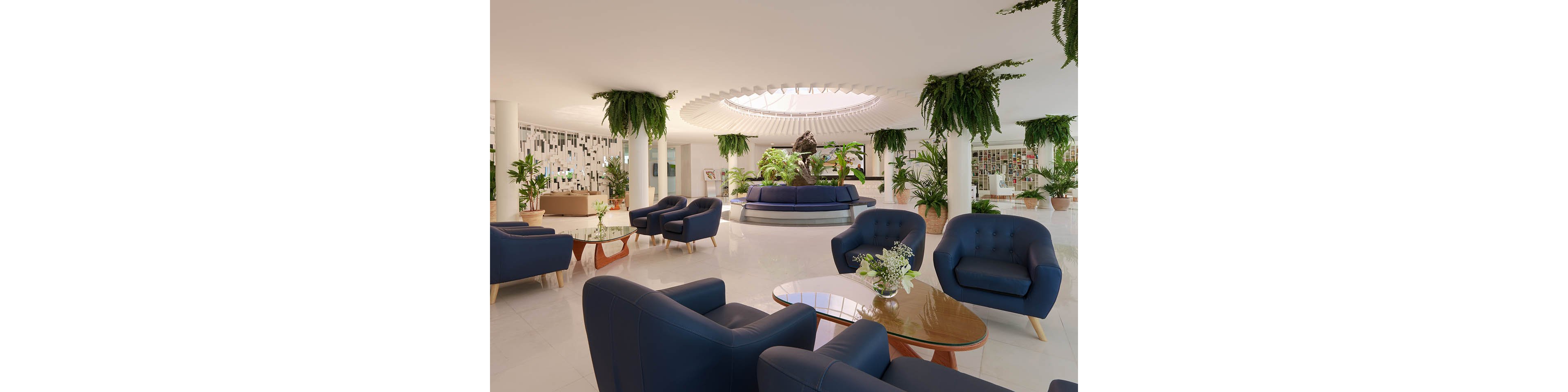 Spacious contemporary lobby features clusters of blue armchairs and glass-topped coffee tables. A circular seating area surrounds a central sculpture beneath a large skylight. Hanging planters and potted greenery add a lush, biophilic touch to the bright white interior. The space appears calm and inviting, suitable for hotel guests or business visitors.