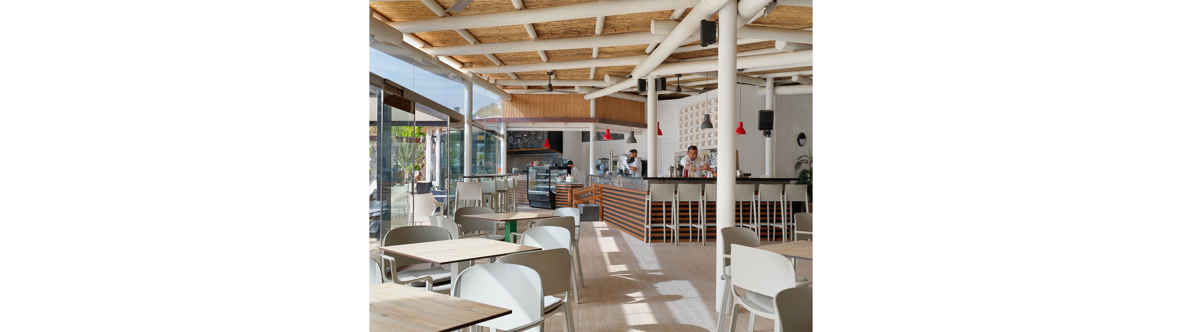 A contemporary café interior features light wooden tables and white chairs arranged in an open seating area. The bar counter at the back is staffed by baristas preparing drinks and food. Large glass walls reveal an outdoor patio and greenery, filling the space with natural light. Red pendant lights, a wood slat ceiling, and clean minimalist décor create a relaxed, modern atmosphere.