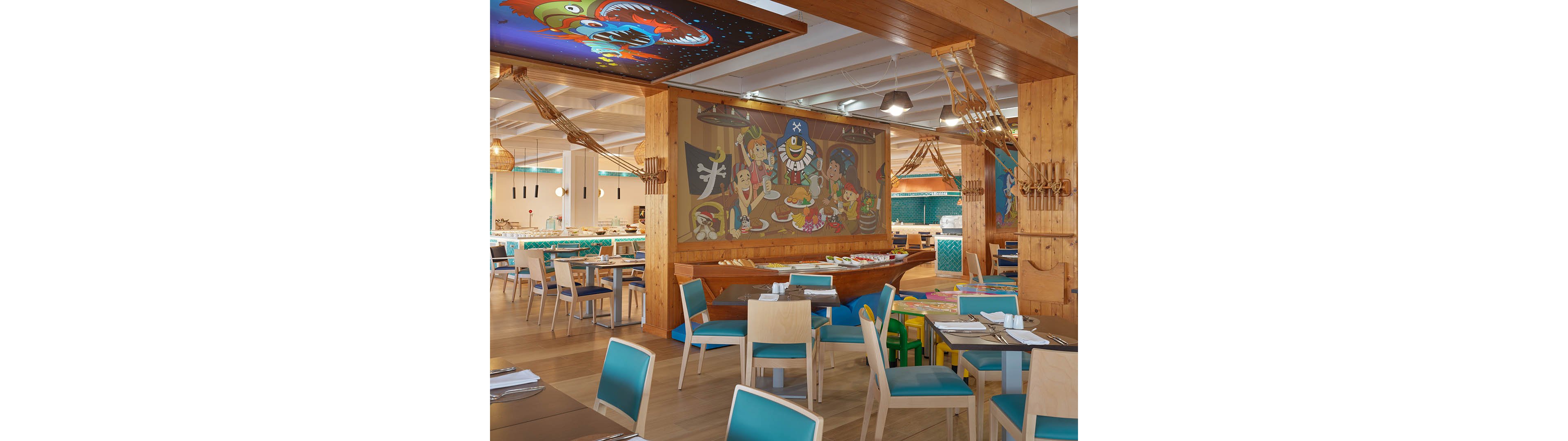 Brightly lit restaurant interior features wooden tables and turquoise chairs arranged neatly. A large cartoon pirate mural dominates the wall, with playful characters enjoying food and drinks. Ceiling artwork and nautical decor elements create a fun, themed dining atmosphere. The space appears family-friendly, with vibrant colors and a casual, welcoming layout.