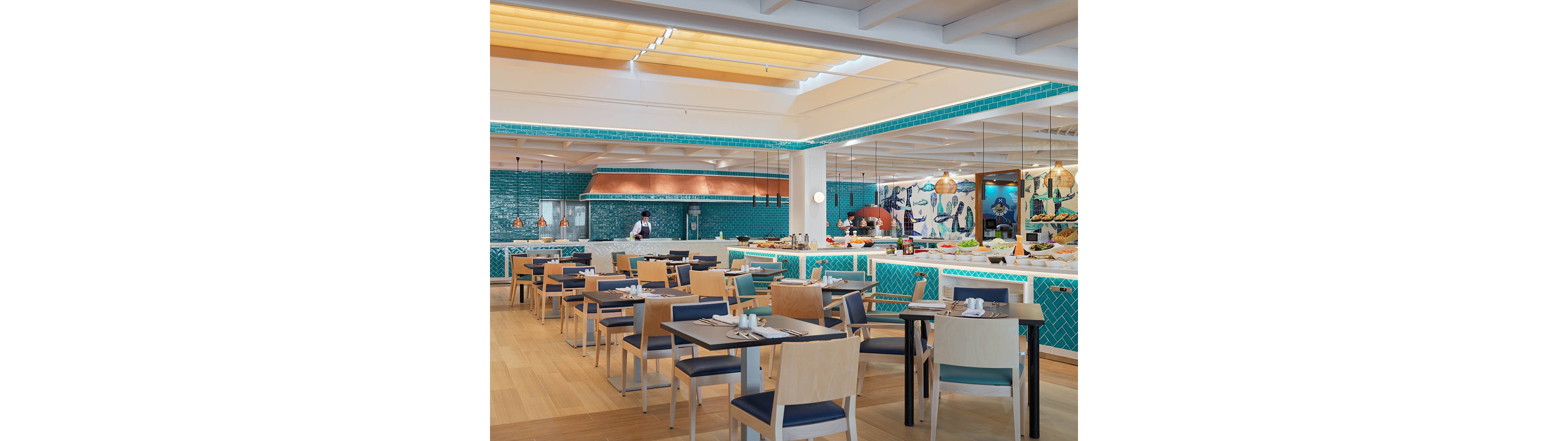 Contemporary restaurant dining room with neatly arranged tables and chairs is shown in a bright, open space. A chef works behind a long counter with turquoise tile accents and an open kitchen area. Warm wooden flooring, pendant lights, and decorative wall art add visual interest and contrast to the cool blue palette. The setting appears clean, inviting, and ready for guests, with table settings prepared for service.