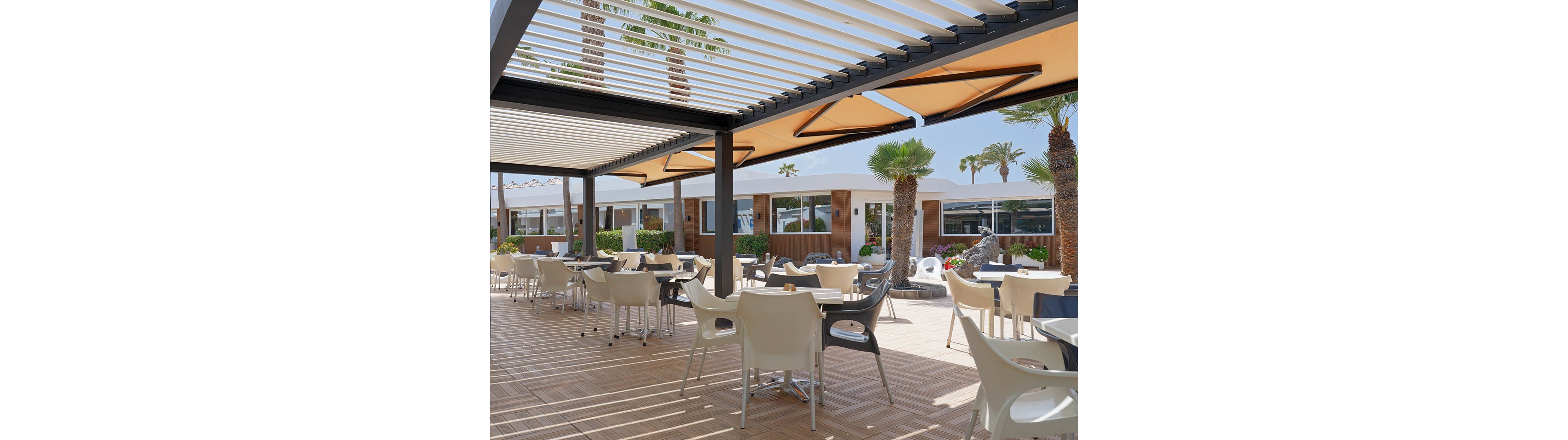 An open-air terrace at a resort features numerous tables and chairs arranged on a wooden deck. A modern pergola with slatted roof panels provides partial shade while allowing sunlight to filter through. Palm trees and landscaped greenery surround the seating area, with low buildings and large windows in the background. The scene suggests a relaxed, vacation atmosphere in a warm, sunny climate.