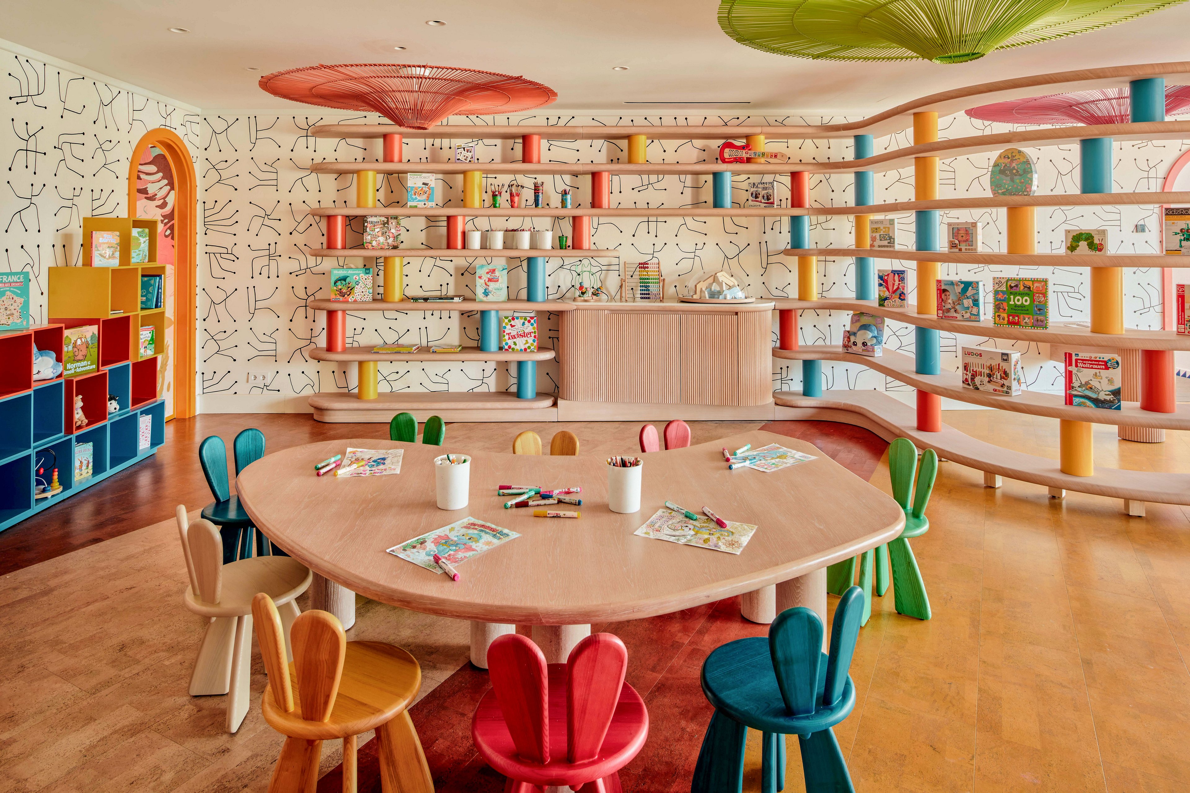 A bright children’s playroom features a large round wooden table surrounded by small colorful chairs. The walls are lined with curved shelves holding books, toys, and games, set against a playful patterned wallpaper. Open cubby storage and ceiling-mounted circular fixtures add to the whimsical design. Activity sheets and drawing supplies on the table suggest a creative, educational environment for kids.