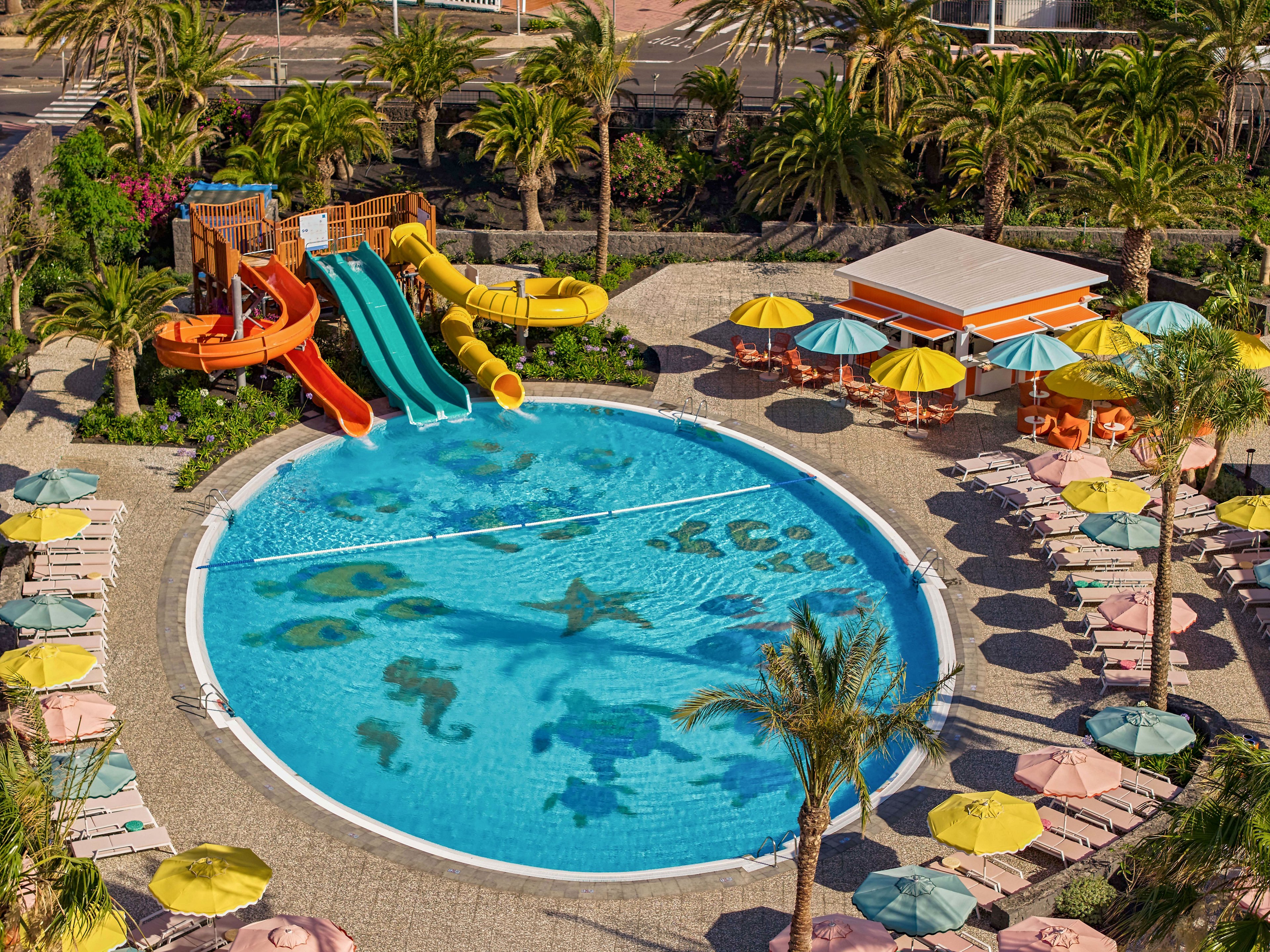 A circular resort swimming pool features playful sea creature mosaics on the pool floor. Multiple bright water slides and a wooden play structure stand at one side of the pool. Yellow umbrellas shade lounge chairs arranged neatly around the pool deck. Palm trees and lush greenery surround the area, creating a tropical vacation atmosphere.