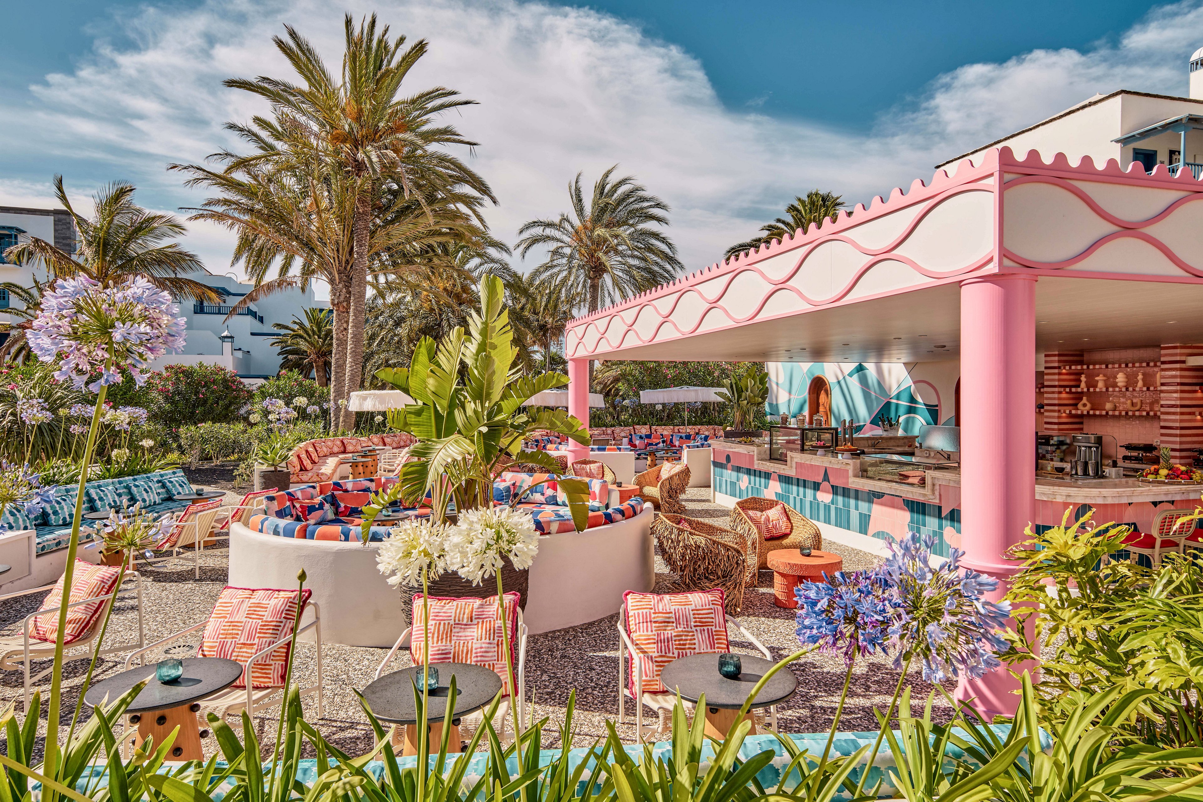 Vibrant outdoor lounge and bar area features colorful seating and bold geometric patterns. The setting is a tropical resort garden with tall palm trees and lush greenery surrounding the space. A pink and white bar structure anchors the scene, complemented by patterned cushions, umbrellas, and decorative plants. The atmosphere feels playful and summery, ideal for relaxing, socializing, and enjoying drinks outdoors.