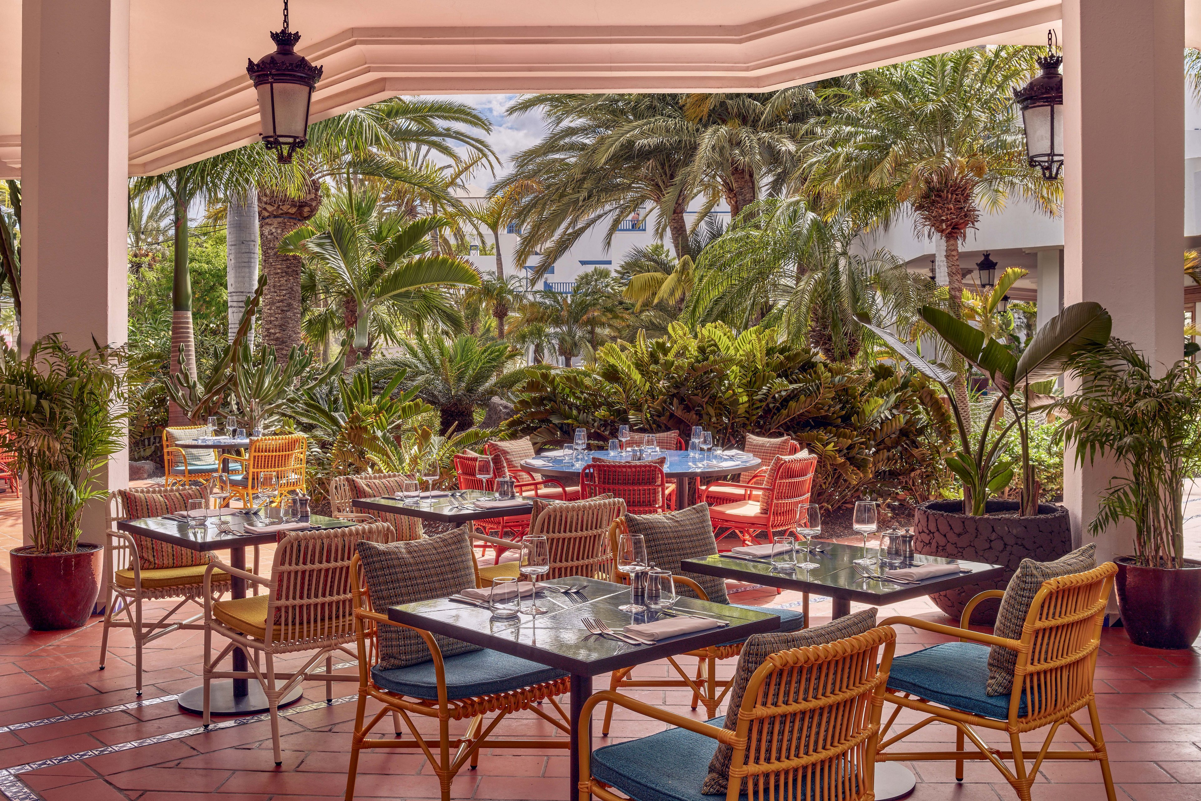 An inviting outdoor restaurant terrace features wicker chairs and glass-topped tables arranged under a covered patio. The setting overlooks lush tropical greenery with palm trees and dense foliage creating a garden atmosphere. Colorful chairs in yellow, red, and blue add vibrancy to the scene, while table settings with glasses and cutlery suggest readiness for guests. Warm terracotta floor tiles and hanging lanterns contribute to a relaxed, vacation-style dining experience.