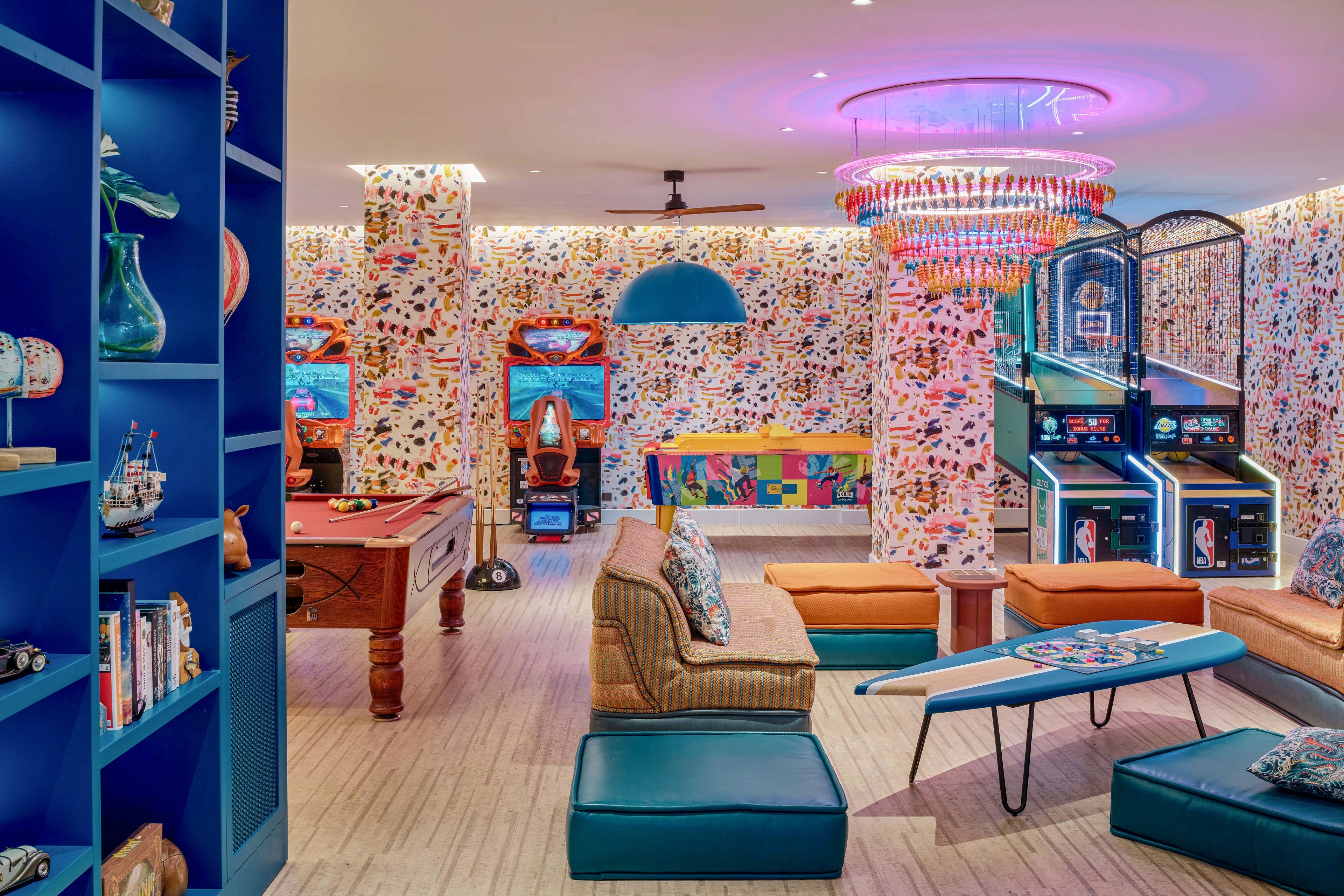 Vibrant lounge space featuring arcade machines, a pool table, and casual seating. The room is decorated with colorful patterned walls and a striking multicolored chandelier that casts a pink glow. A blue shelving unit with books and decorative objects lines one side, while cushioned benches and ottomans create a relaxed hangout area. The overall atmosphere is playful and retro, designed for socializing and gaming.
