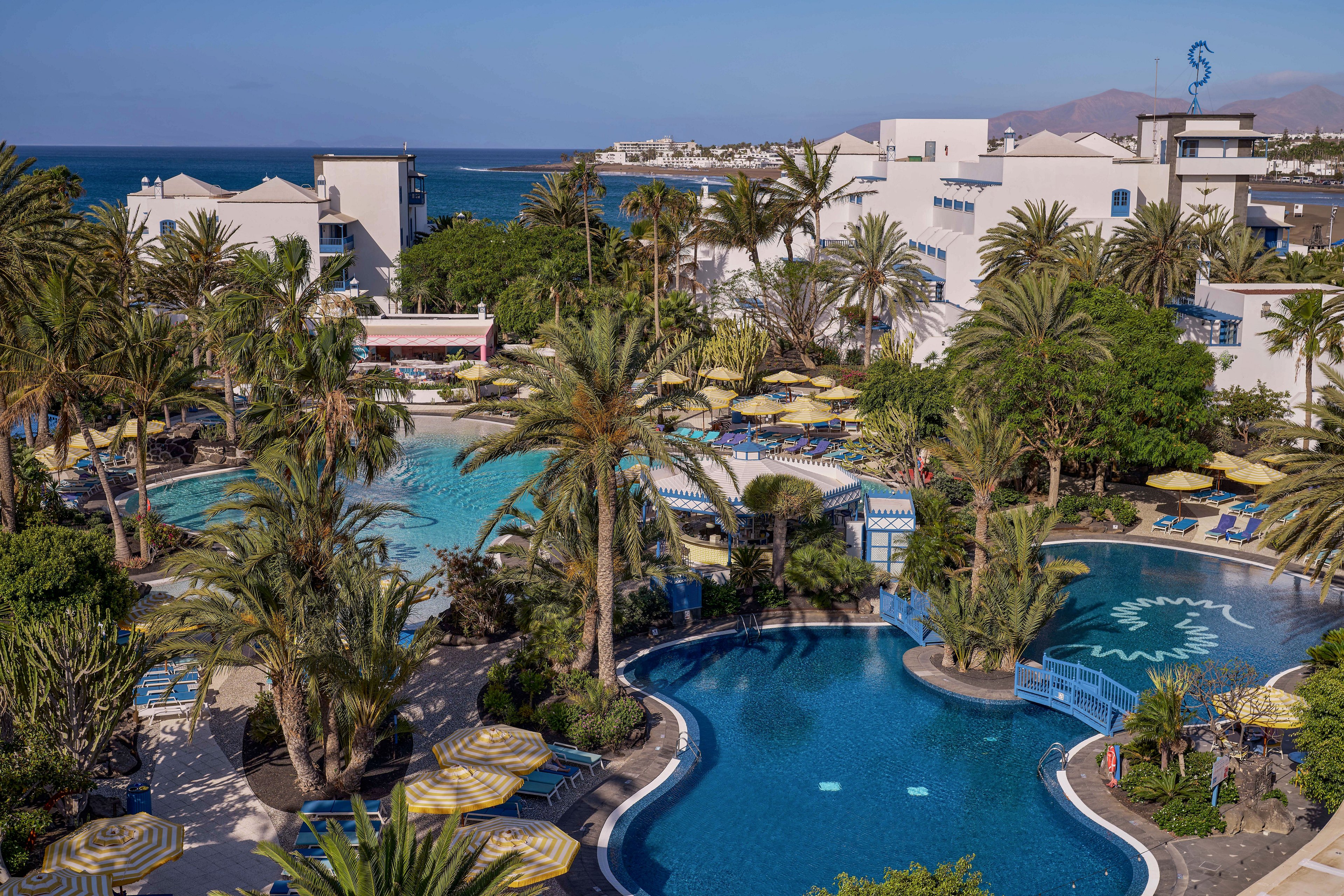 A large seaside resort features multiple outdoor swimming pools surrounded by lush palm trees. Sun loungers with striped yellow umbrellas line the poolside terraces, creating a relaxed vacation atmosphere. White hotel buildings rise behind the pools, with views extending to the blue ocean and distant coastline. The scene suggests a warm, sunny day ideal for leisure, swimming, and sunbathing.