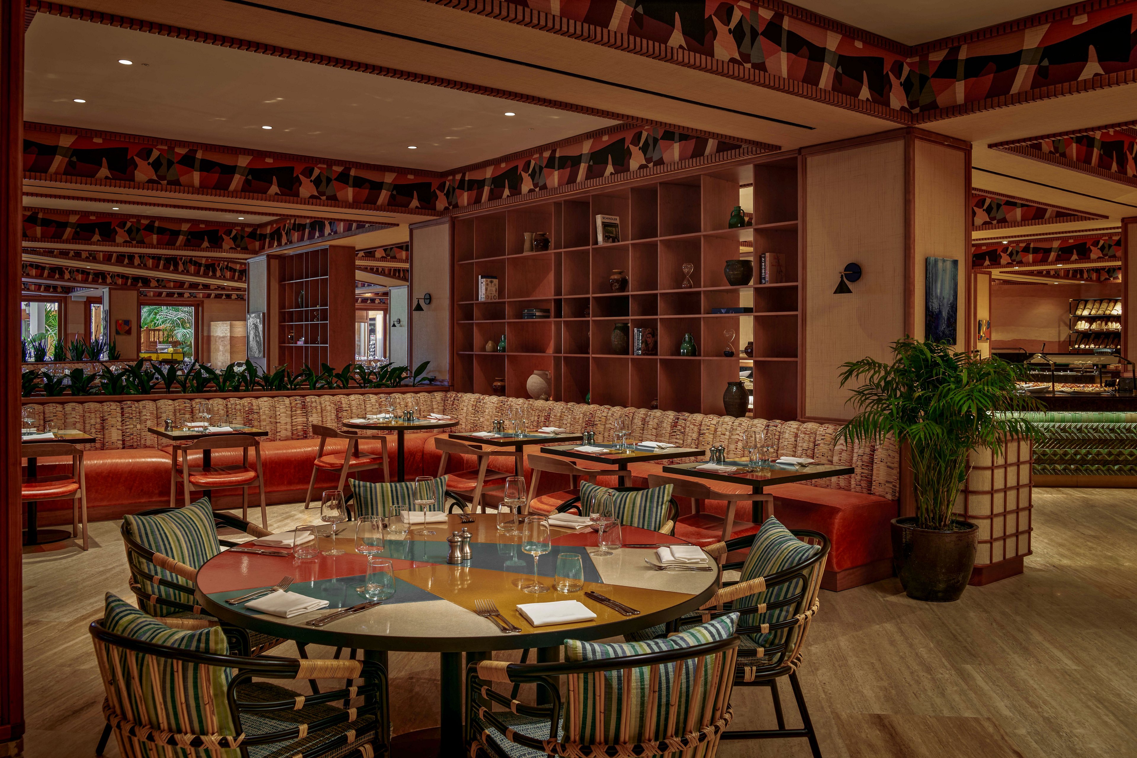 A warmly lit restaurant interior features round and rectangular tables arranged with neatly set place settings. Patterned upholstered chairs and cushioned banquette seating surround the tables, creating a cozy and inviting dining area. Decorative shelving with vases and objects lines the back wall, while plants add greenery to the space. The overall design combines modern furnishings with colorful geometric accents for a sophisticated atmosphere.