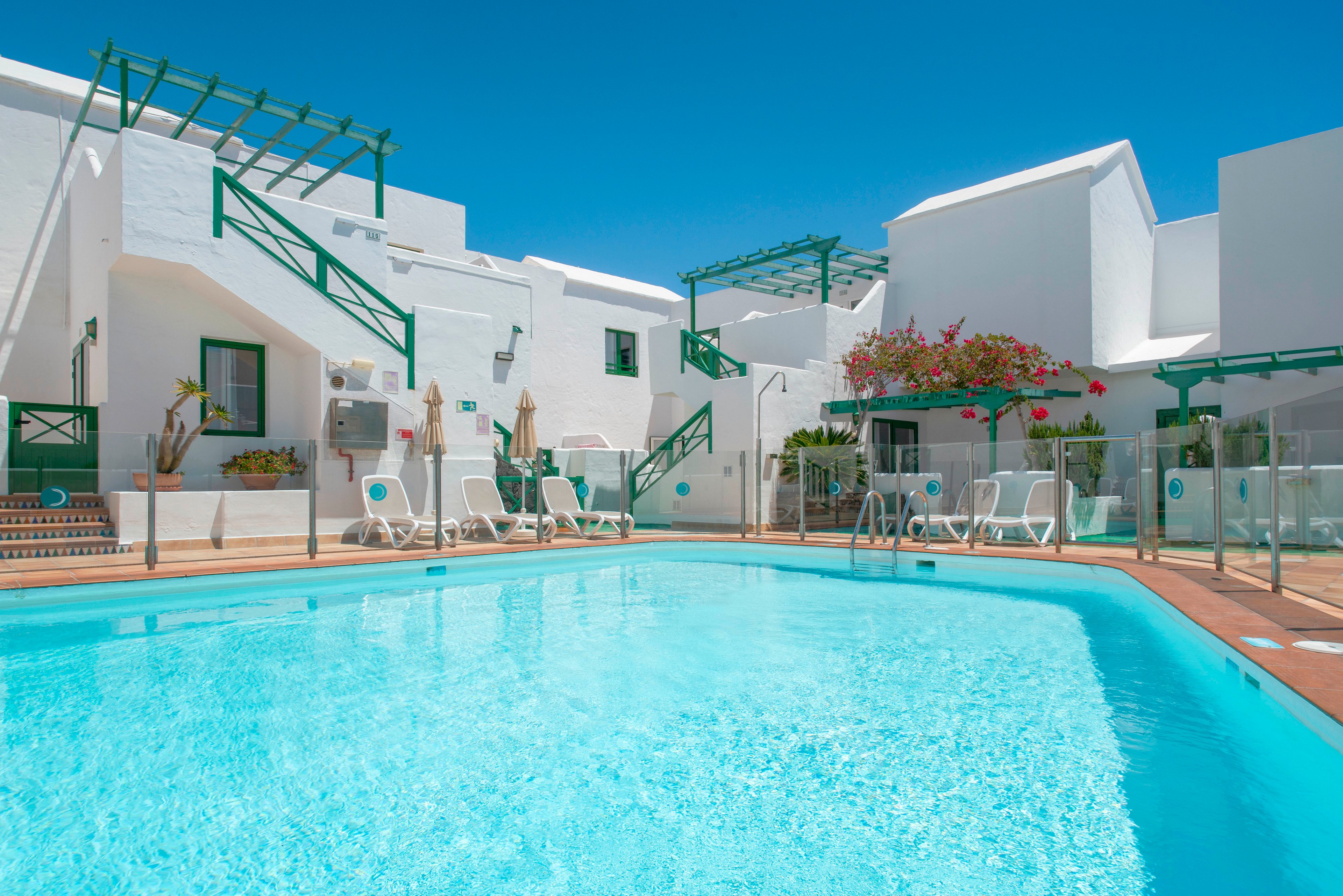 Bright white resort buildings surround a clear blue outdoor swimming pool under a cloudless sky. Green railings and pergolas add contrast to the minimalist architecture. Sun loungers and umbrellas line the poolside deck, suggesting a relaxing holiday setting. Flowering plants and glass safety barriers frame the water, enhancing the clean and inviting atmosphere.