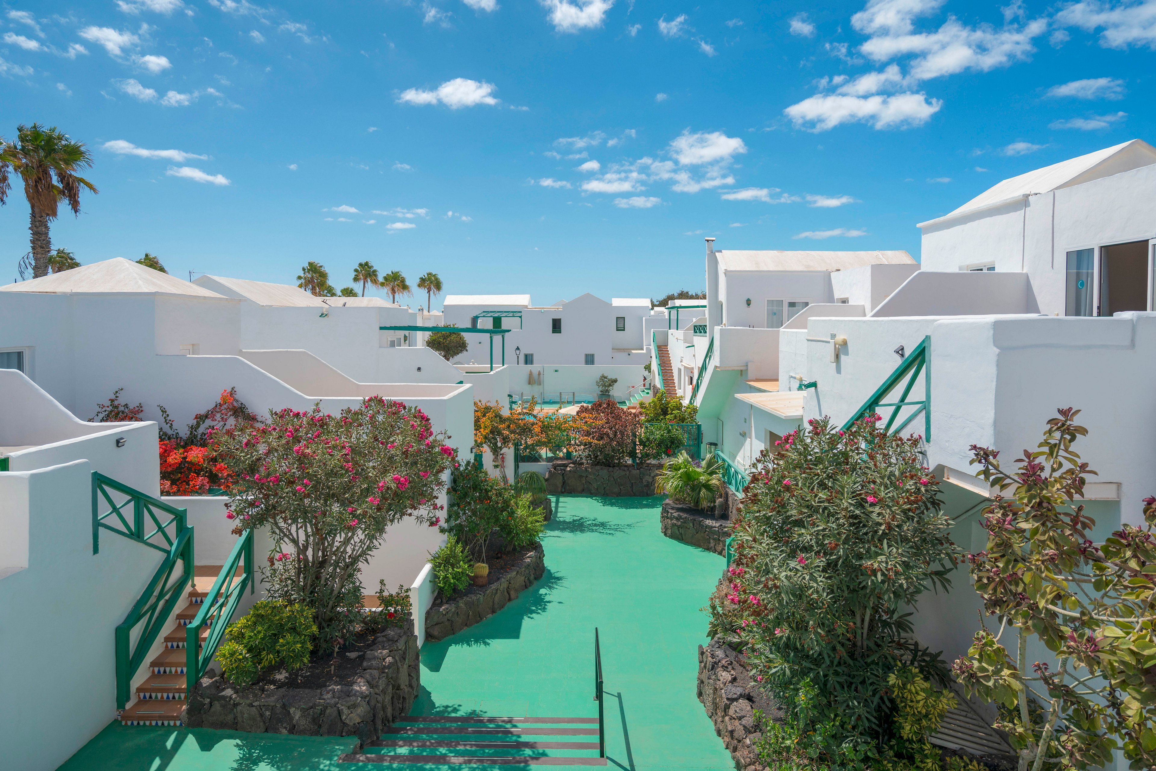 A bright white resort complex is shown under a clear blue sky with scattered clouds. The scene features a central green walkway lined with flowering shrubs and tropical plants. White cubic buildings with green railings and accents create a clean, modern Mediterranean feel. Palm trees rise in the background, enhancing the relaxed holiday atmosphere.