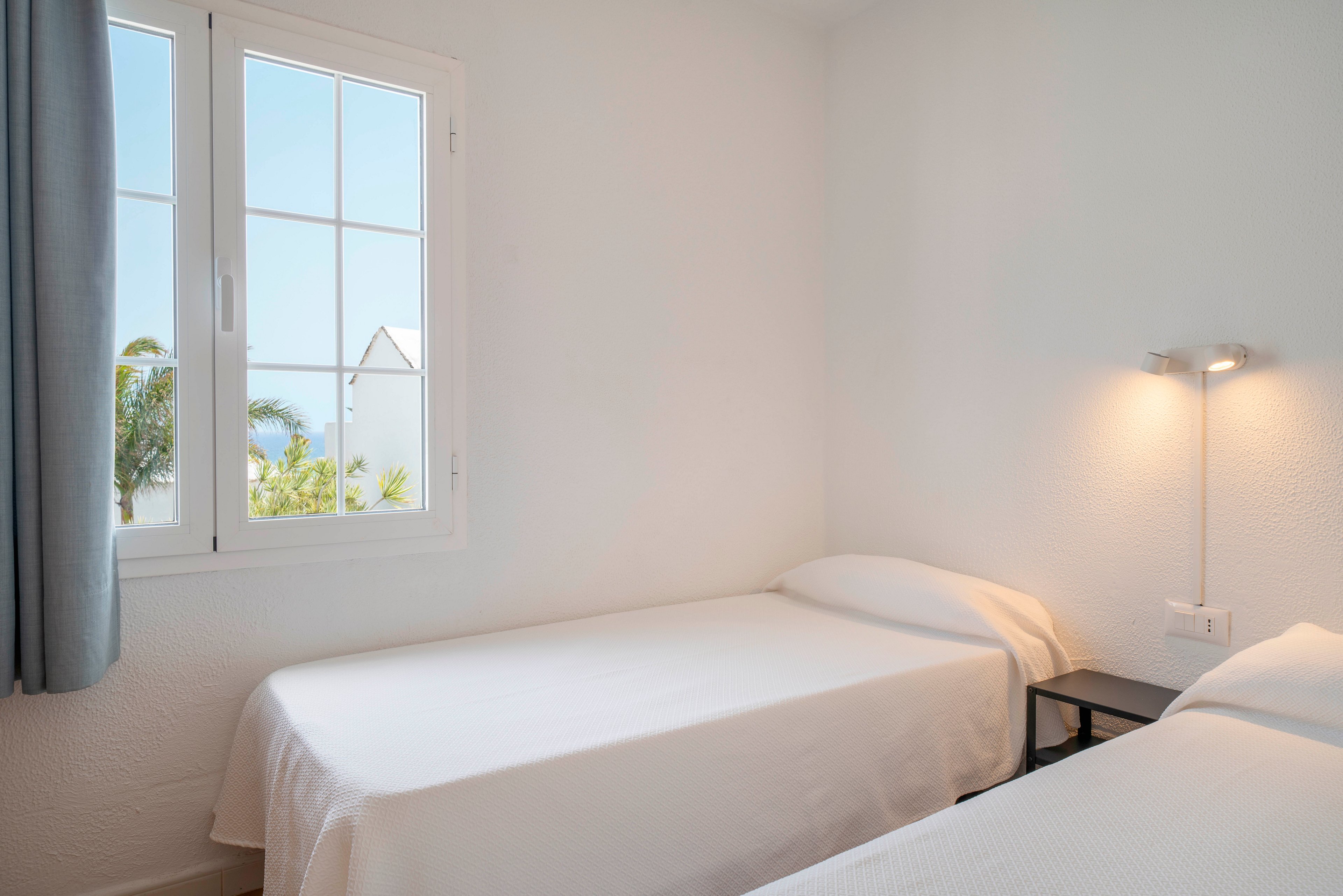 A simple twin bedroom features two neatly made single beds with white linens against a textured white wall. Natural light enters through a large window, revealing palm trees and a bright sky outside. A slim wall-mounted reading lamp illuminates the space beside a small black bedside table. The overall scene feels clean, airy, and minimal, emphasizing calm and relaxation.